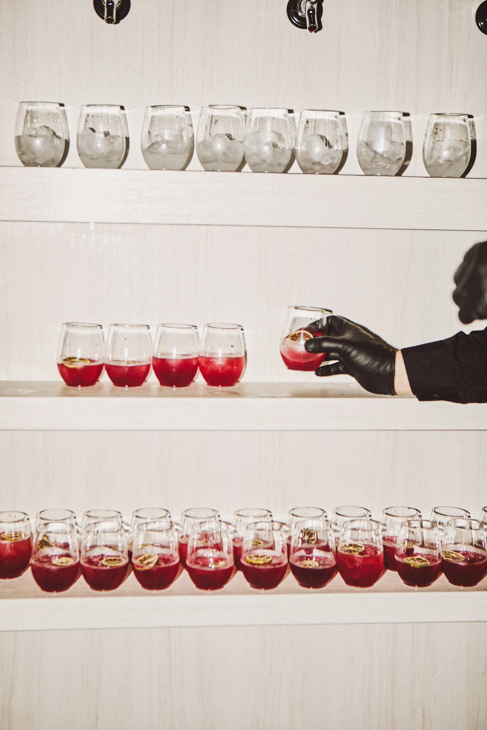 A person wearing black gloves reaching to pick up a glass of red cocktail with lemon slices, standing next to rows of similar glasses filled with red drinks on white shelves.