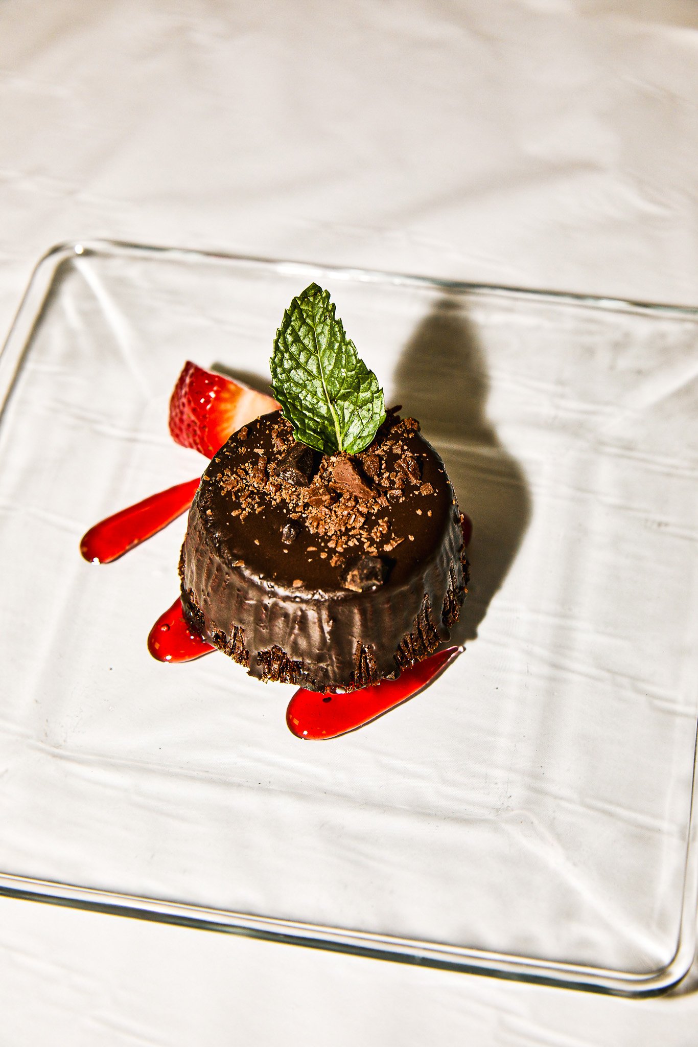 Chocolate dessert garnished with a mint leaf, served with strawberry slices and red sauce on a clear glass plate.