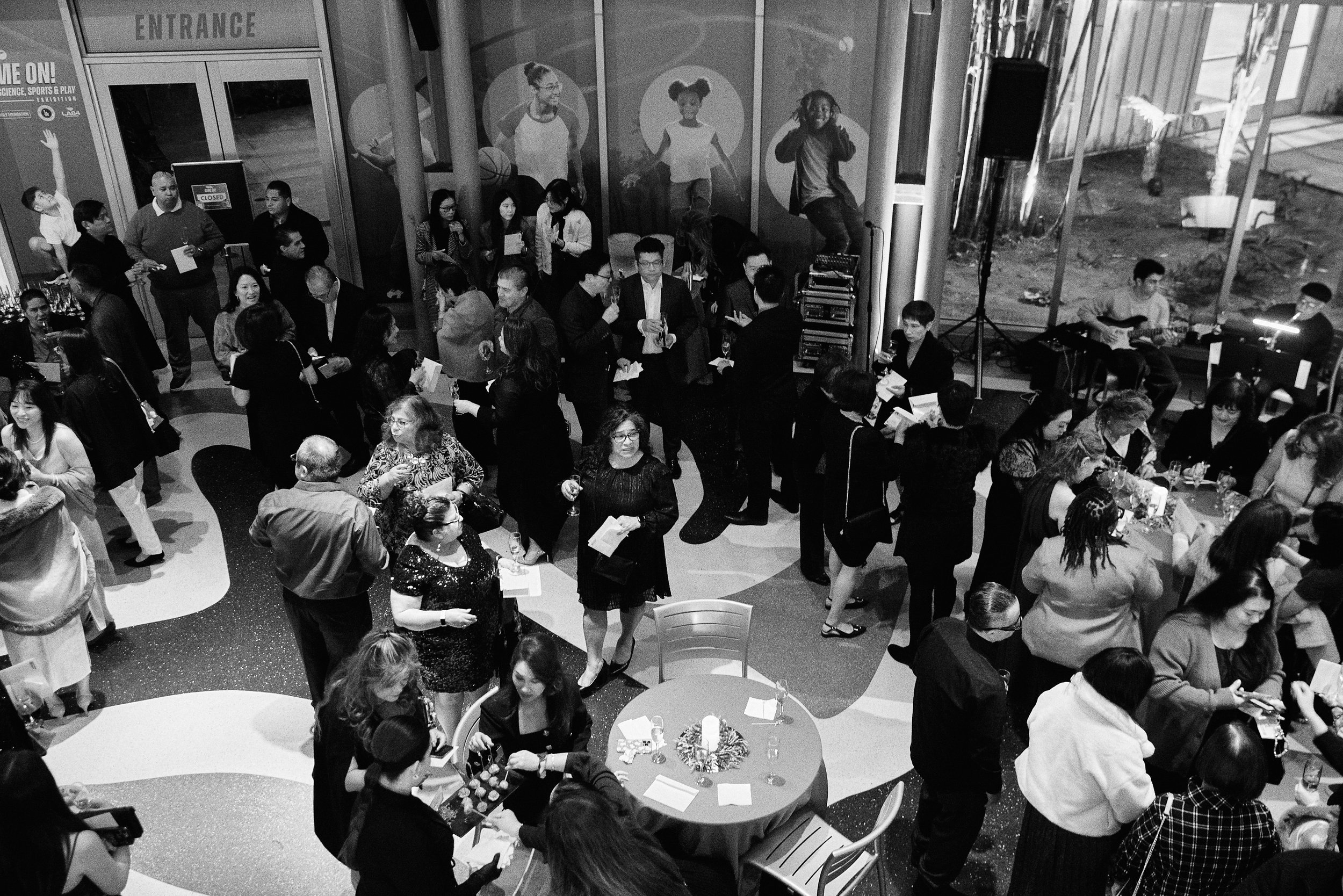 Black and white photo of a crowded indoor reception with people talking and mingling, a musician playing guitar on a stage, and decorative wall art featuring children.