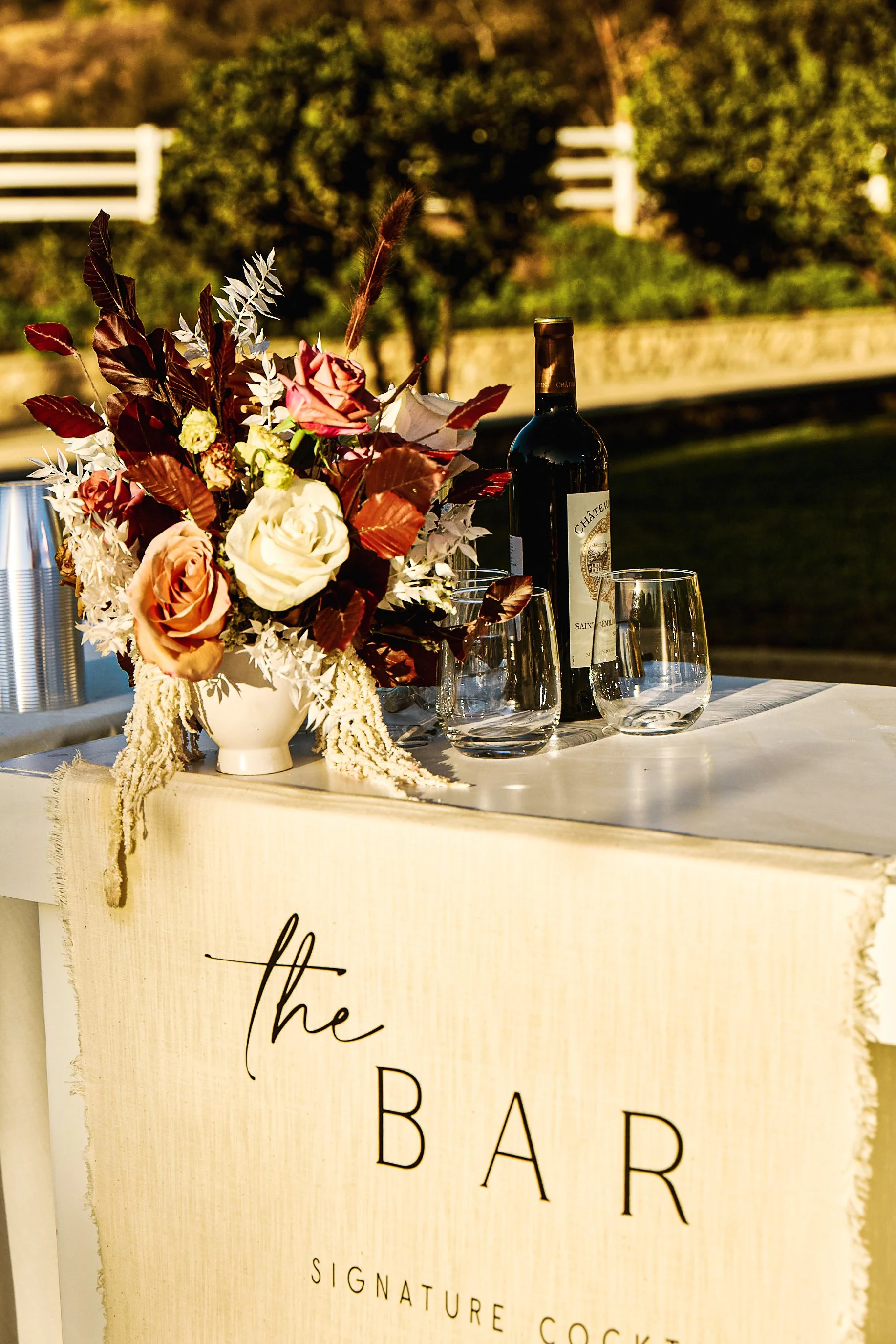 A bar setup with a flower arrangement, a bottle of red wine, and empty glasses, with a sign that reads 'the BAR' in elegant text.