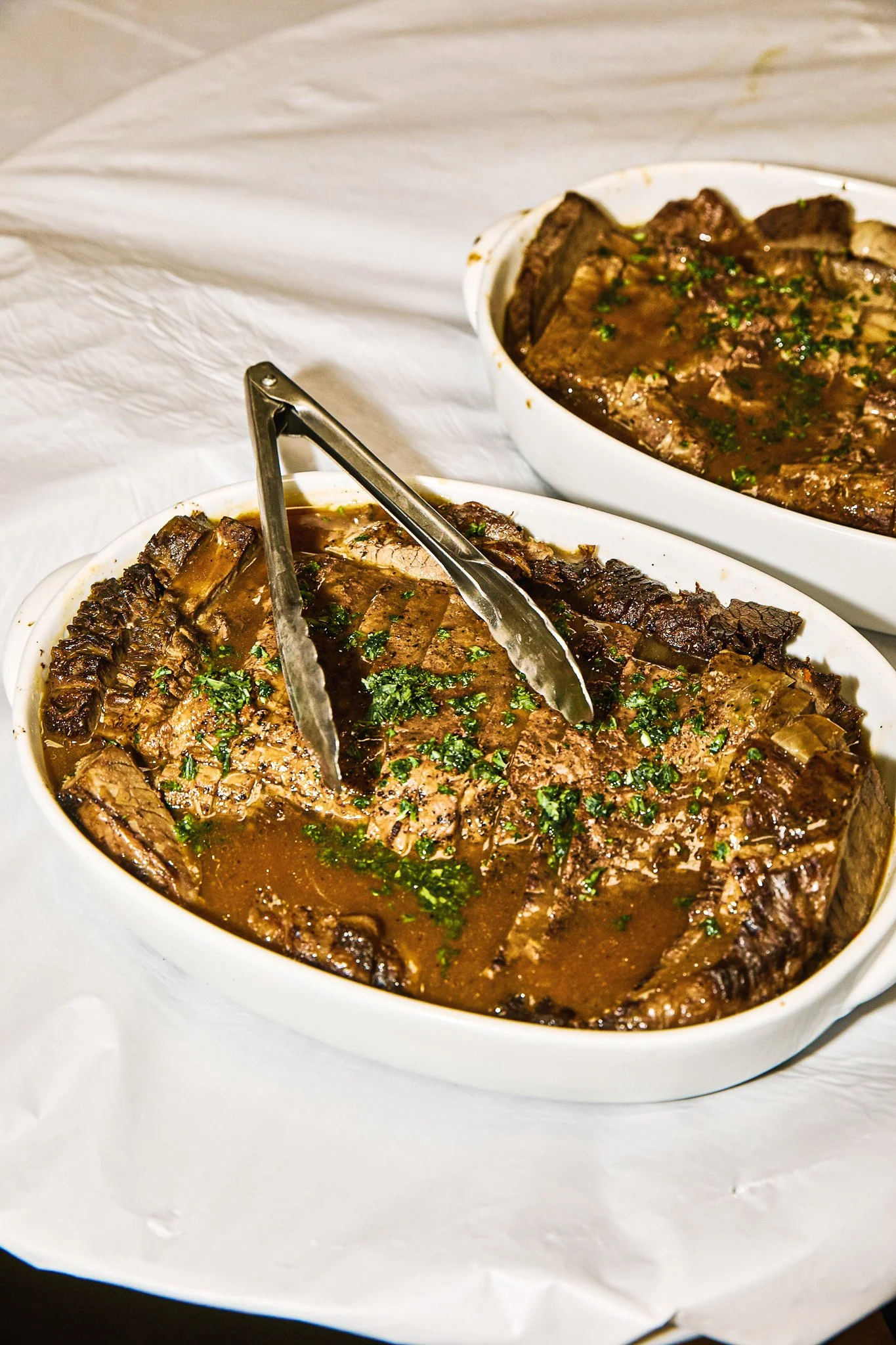 Two white oval dishes filled with braised beef ribs garnished with chopped parsley, placed on a white surface.