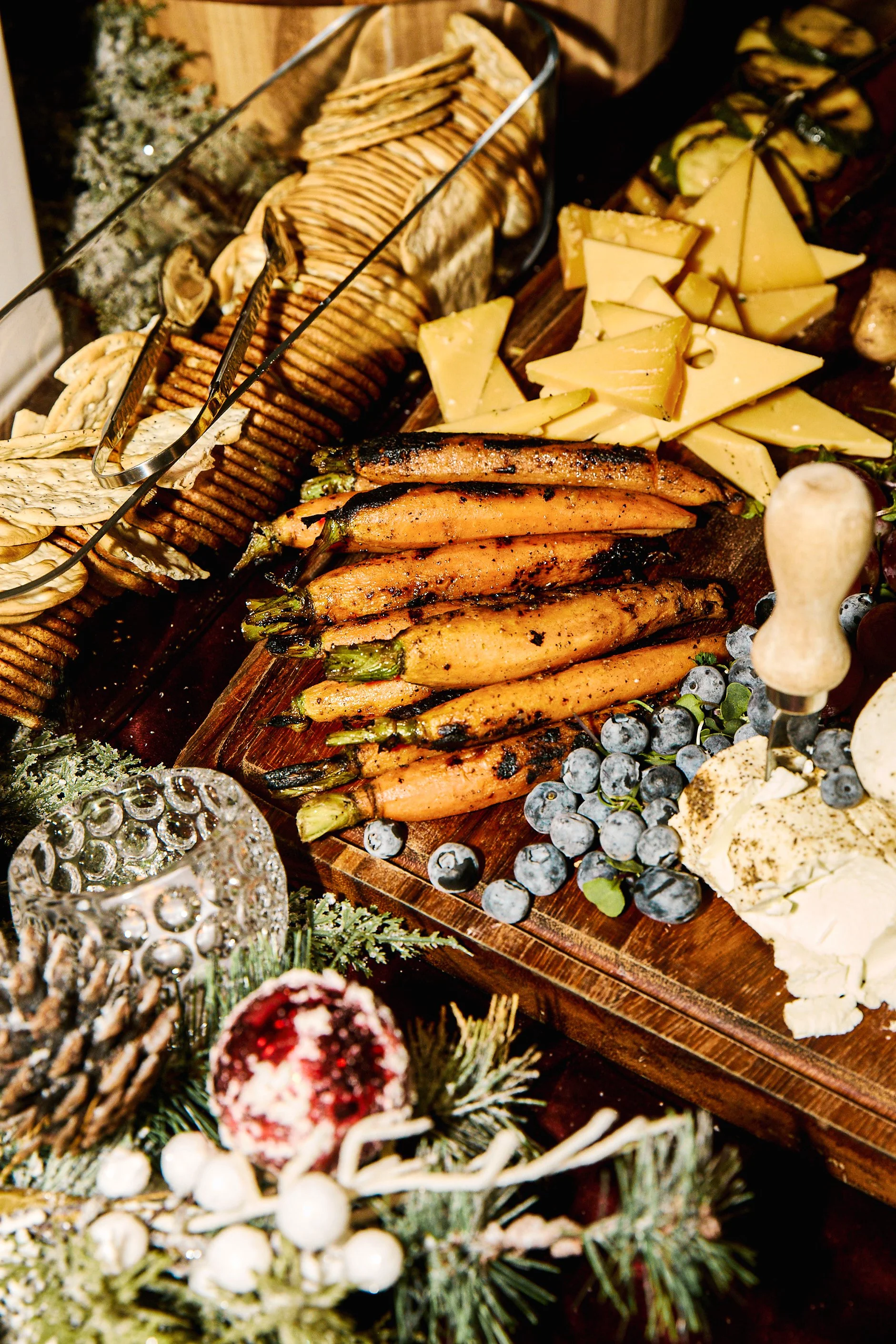 A cheese and charcuterie board featuring slices of cheese, grilled carrots, roasted cherry tomatoes, and crackers, decorated with blueberries and festive greenery.