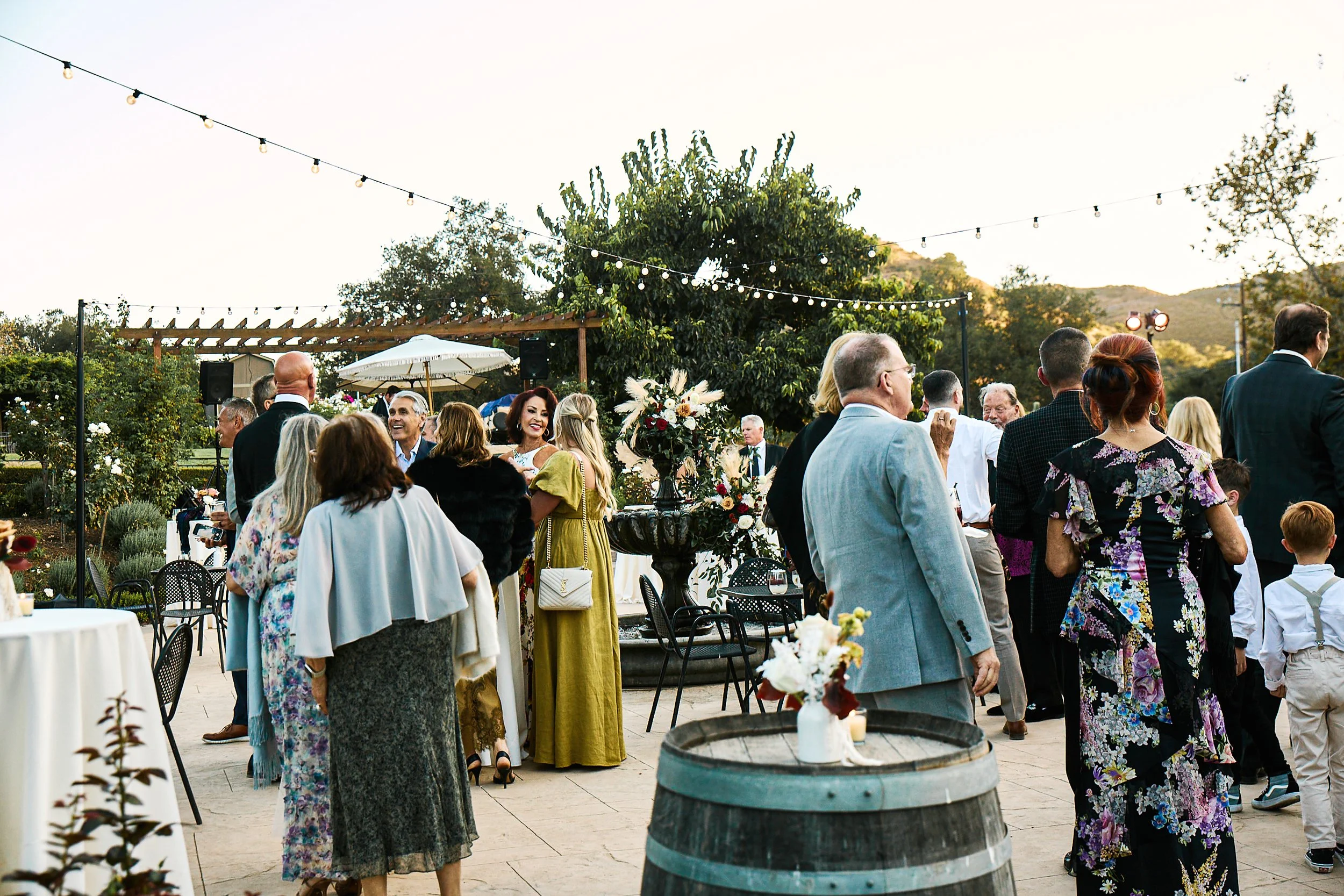 People attending outdoor event, dancing and socializing among decorated tables and floral arrangements under string lights and trees at sunset.