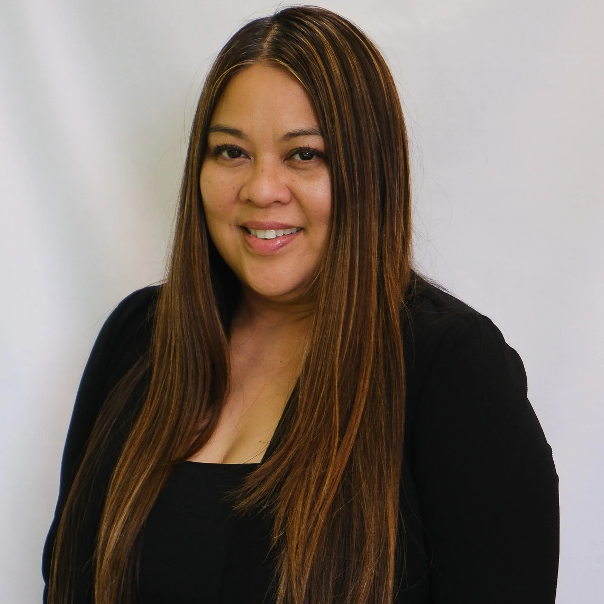 Headshot of a smiling woman with long brown hair, wearing a black blazer and black top against a gray background.