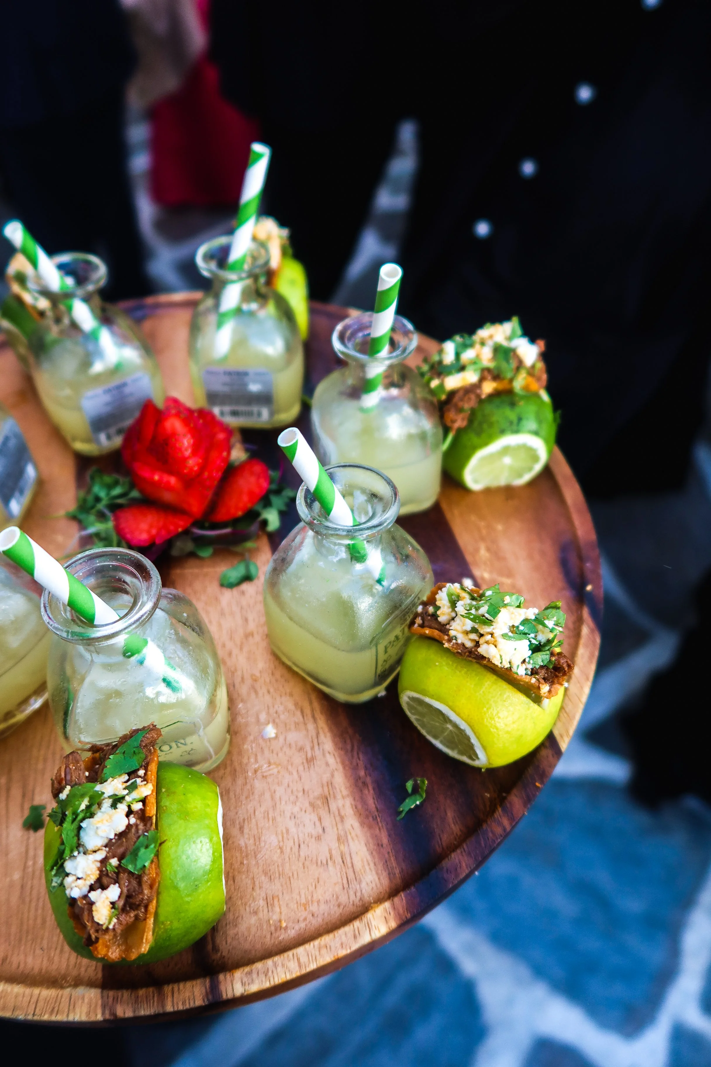 A wooden serving tray holding bottles of lime-infused beverages with green and white striped straws, halved limes with toppings, and fresh strawberries.