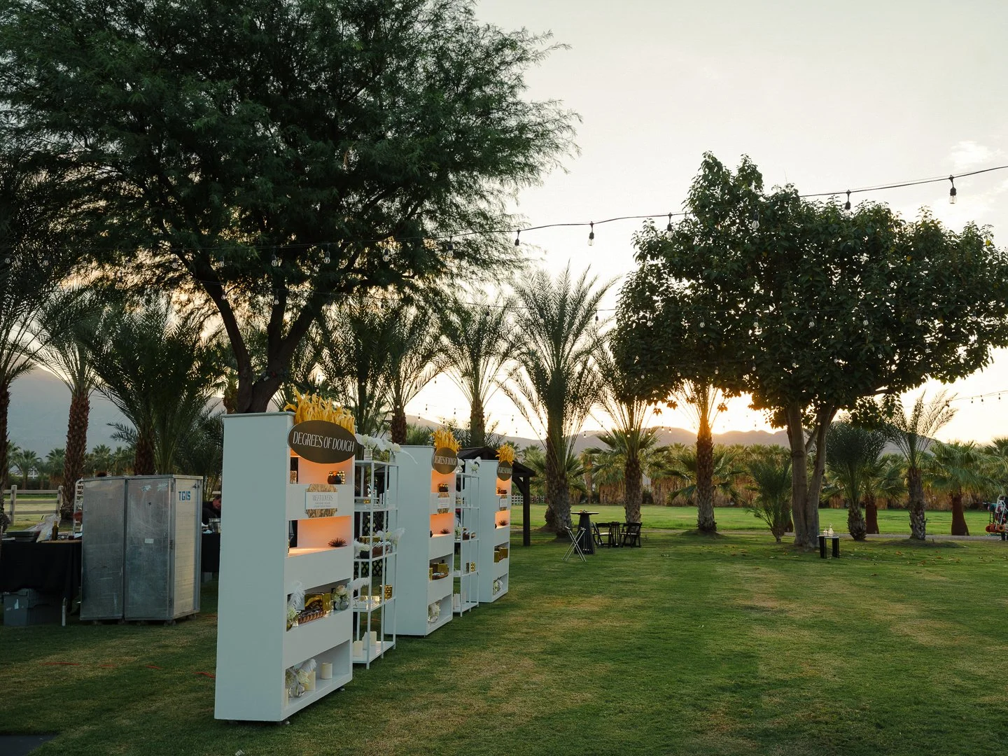 Outdoor event setup on a grassy field with palm trees, standing white shelves labeled "Degrees of Dough" displaying various items, and string lights hanging overhead during sunset.