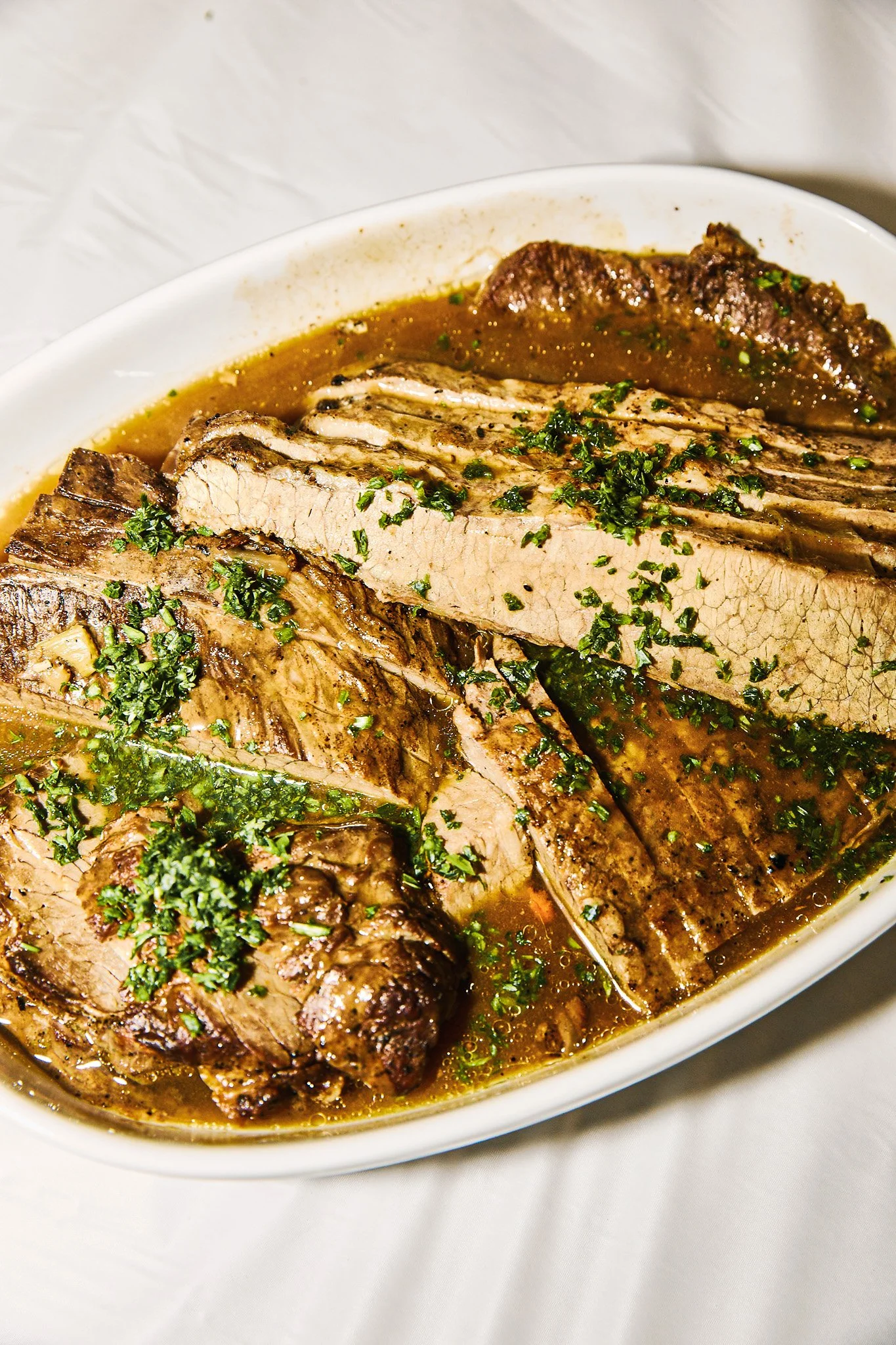 Sliced roast beef with herbs in a gravy inside an oval white dish.