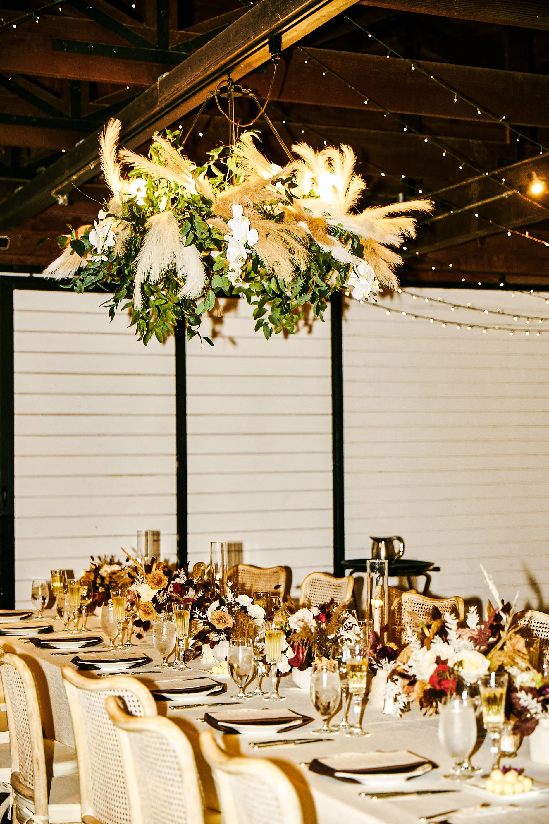 A decorated reception table with floral centerpieces and an overhead floral chandelier, set in a rustic indoor venue with string lights and wooden beams.