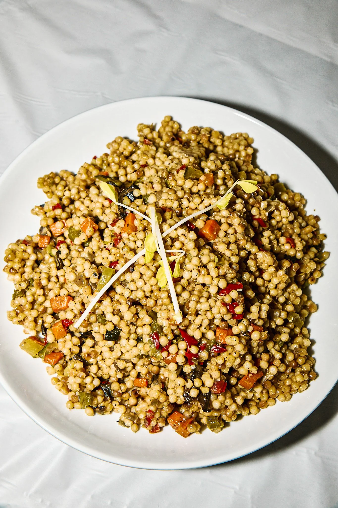 A white plate filled with couscous salad topped with sprouts and colorful chopped vegetables, placed on a white tablecloth.