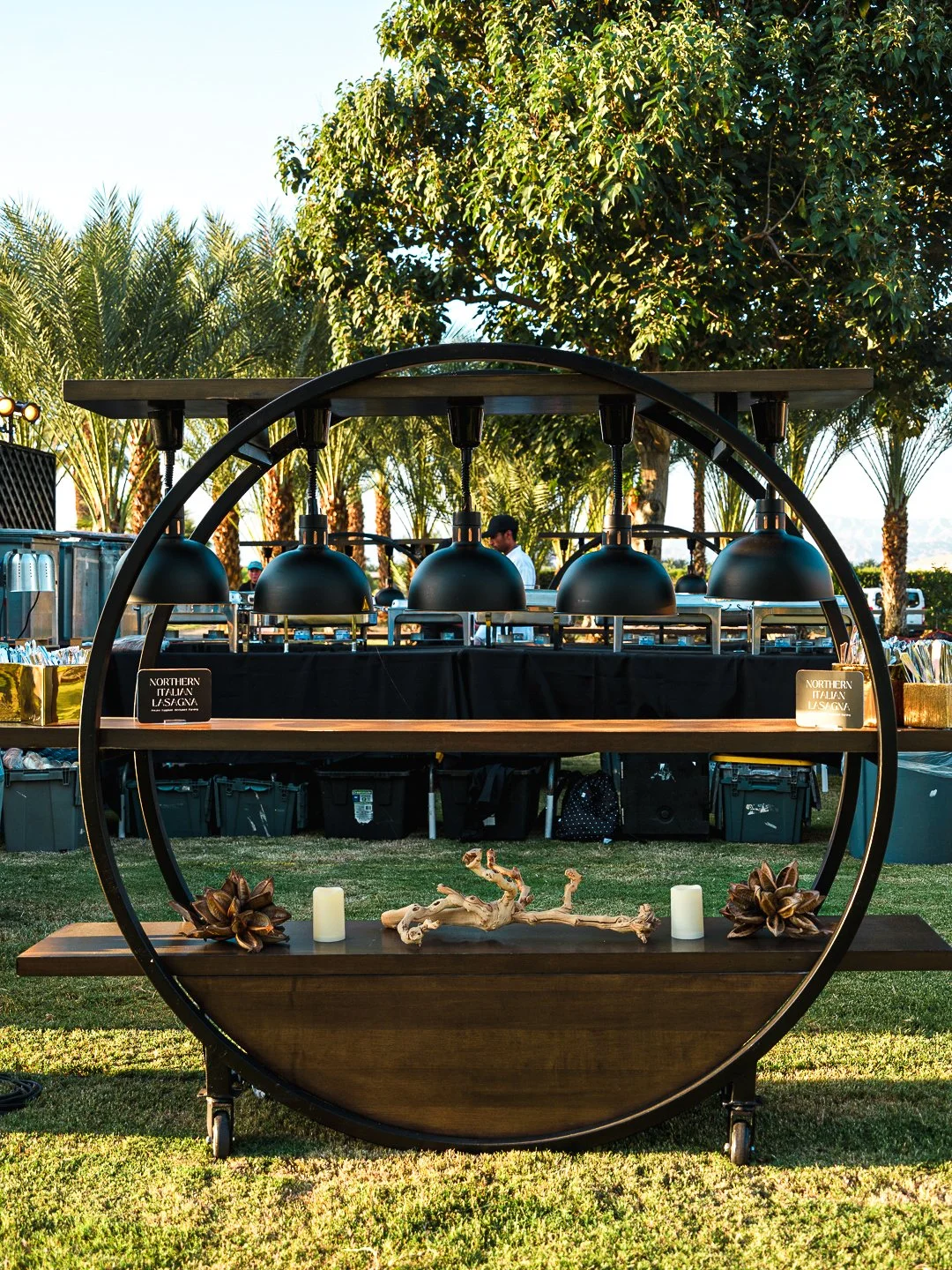 Outdoor buffet station with black pendant lamps, decorative candles, driftwood, and pinecones, set against a background of palm trees and clear sky.