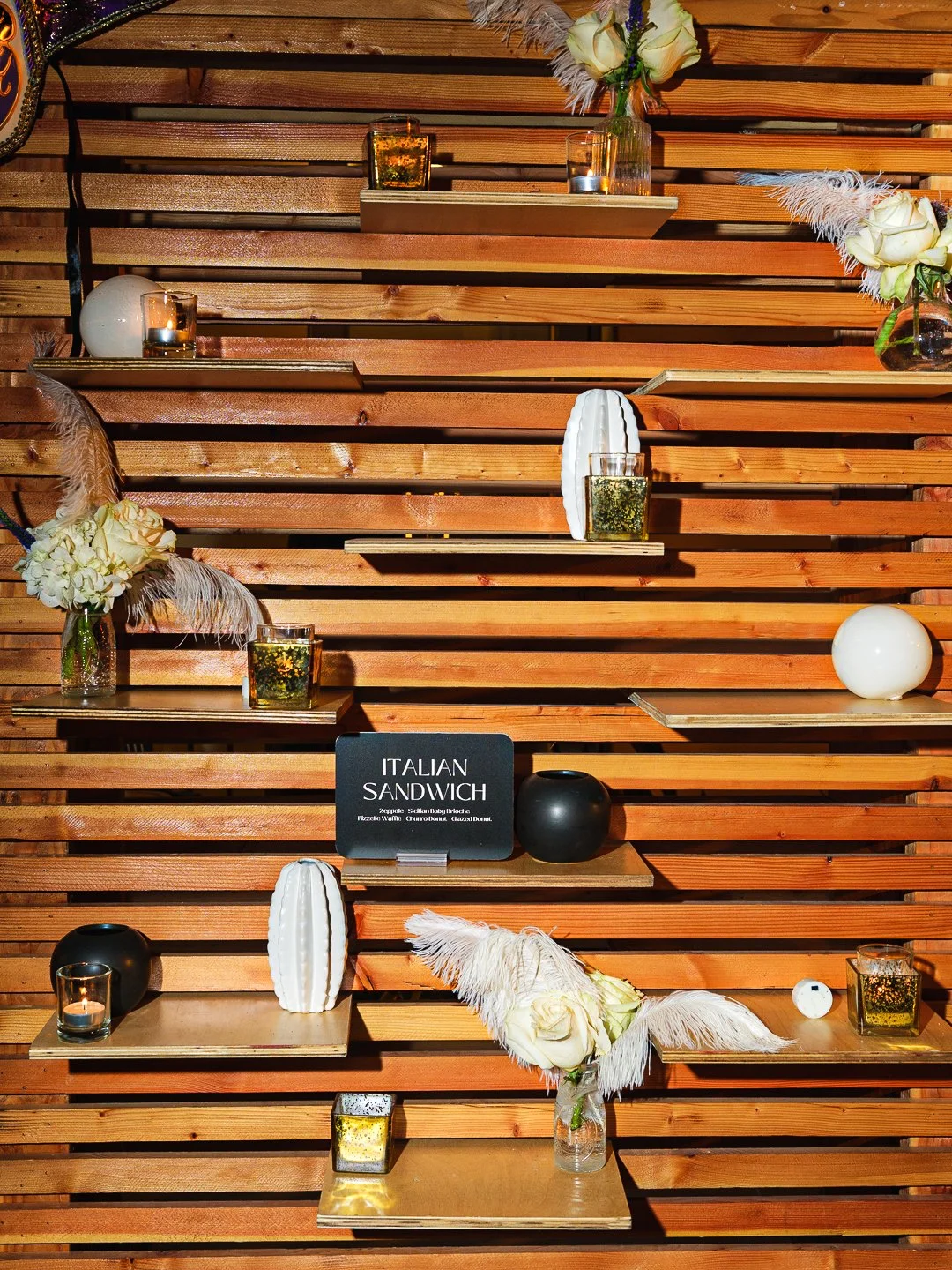 Decorative display with flowers, candles, vases, and a sign for Italian sandwiches on a wooden slat wall.