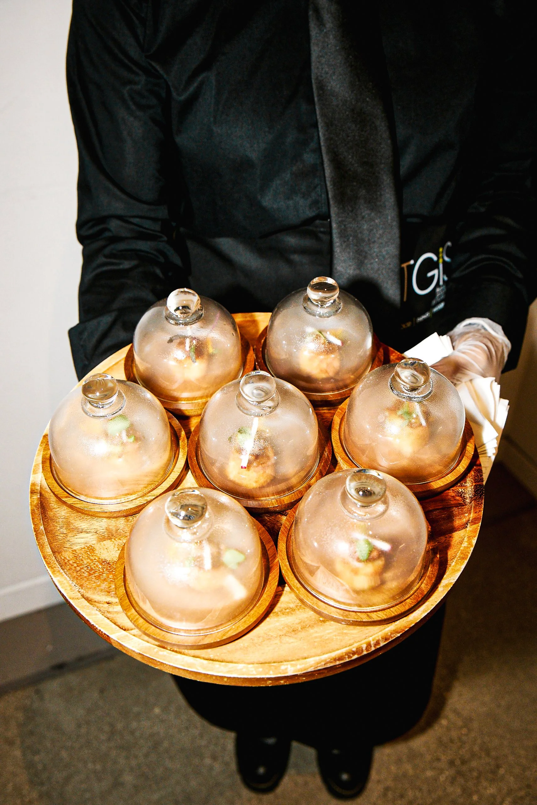 A waiter in black attire and gloves holds a round wooden tray with six glass covered dishes containing food, at an indoor event.