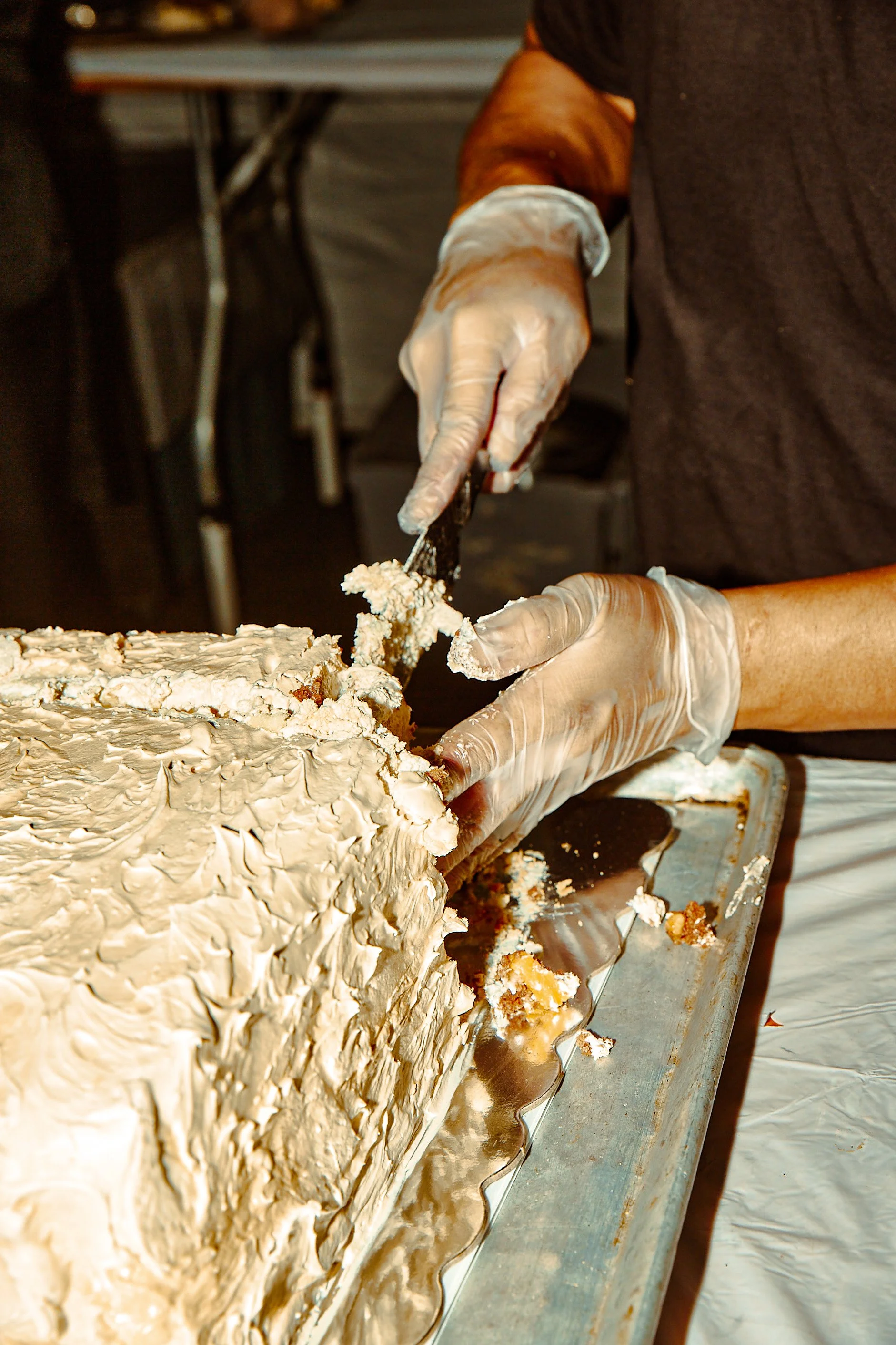 A person wearing gloves slices a large cake covered in white frosting.