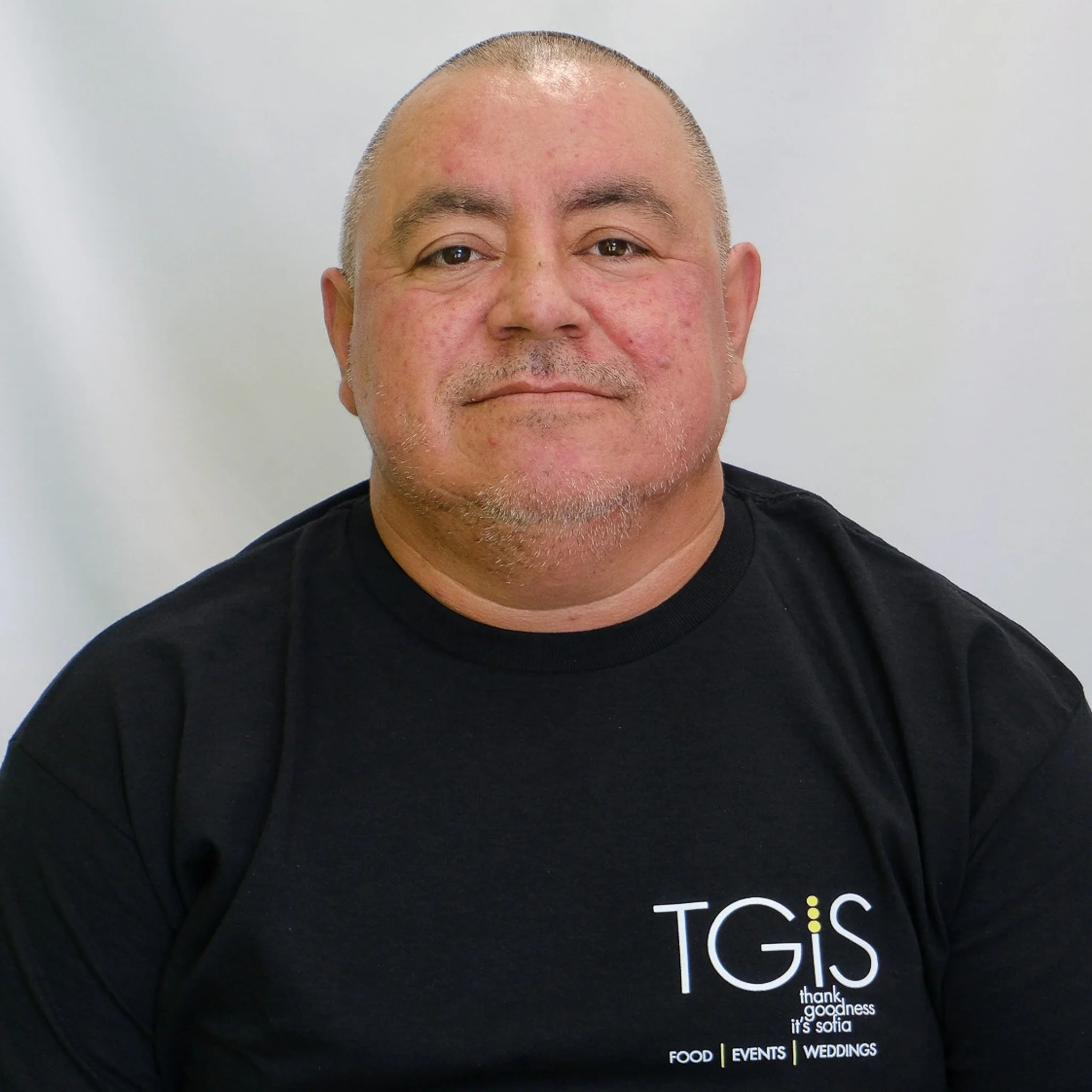 Professional headshot of a smiling man with a shaved head, thick eyebrows, wearing a navy blue collared shirt against a gray background.