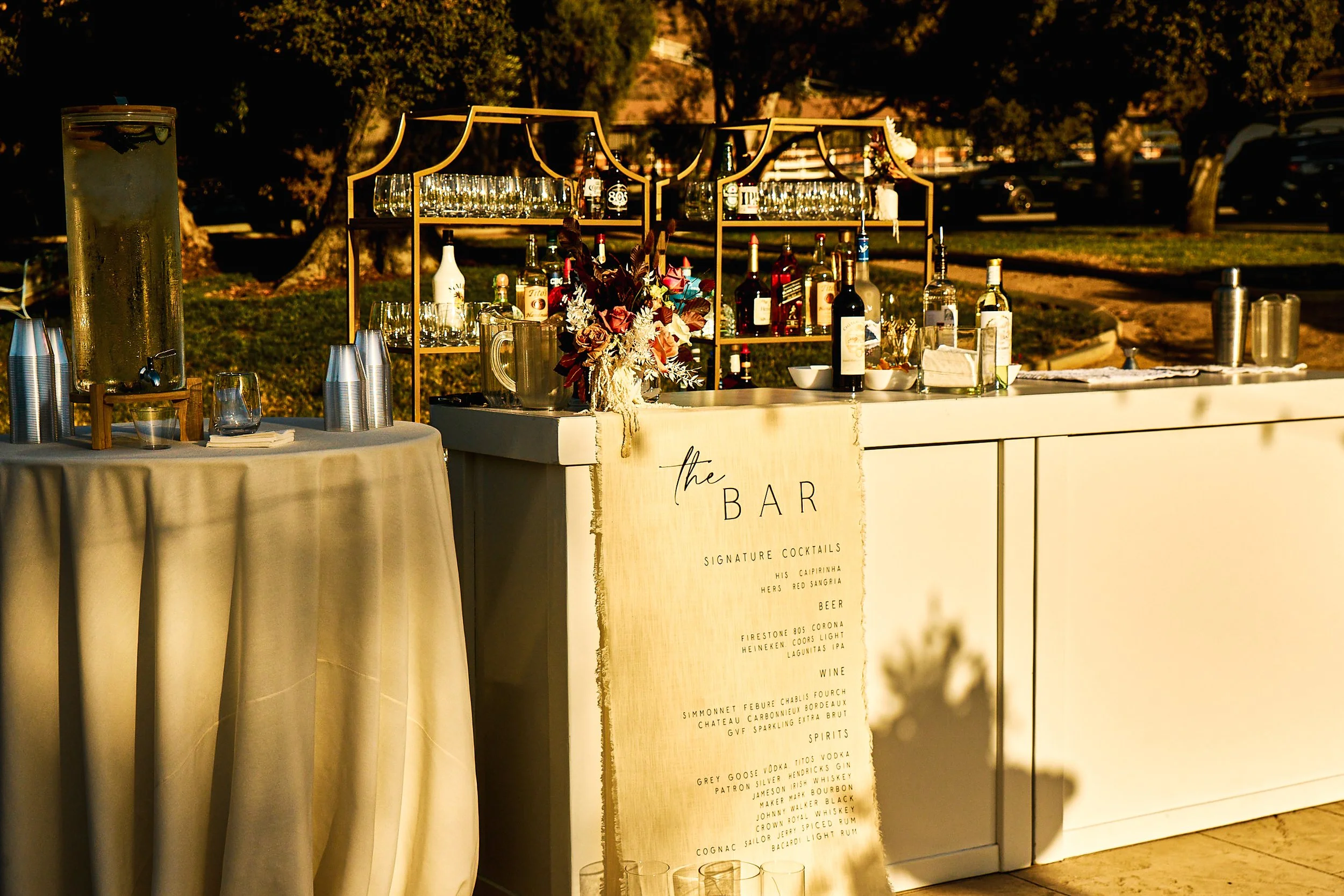 An outdoor bar setup during sunset with a beige sign listing drinks, a floral arrangement, and bottles of alcohol on shelves behind the bar, with trees and parked cars in the background.