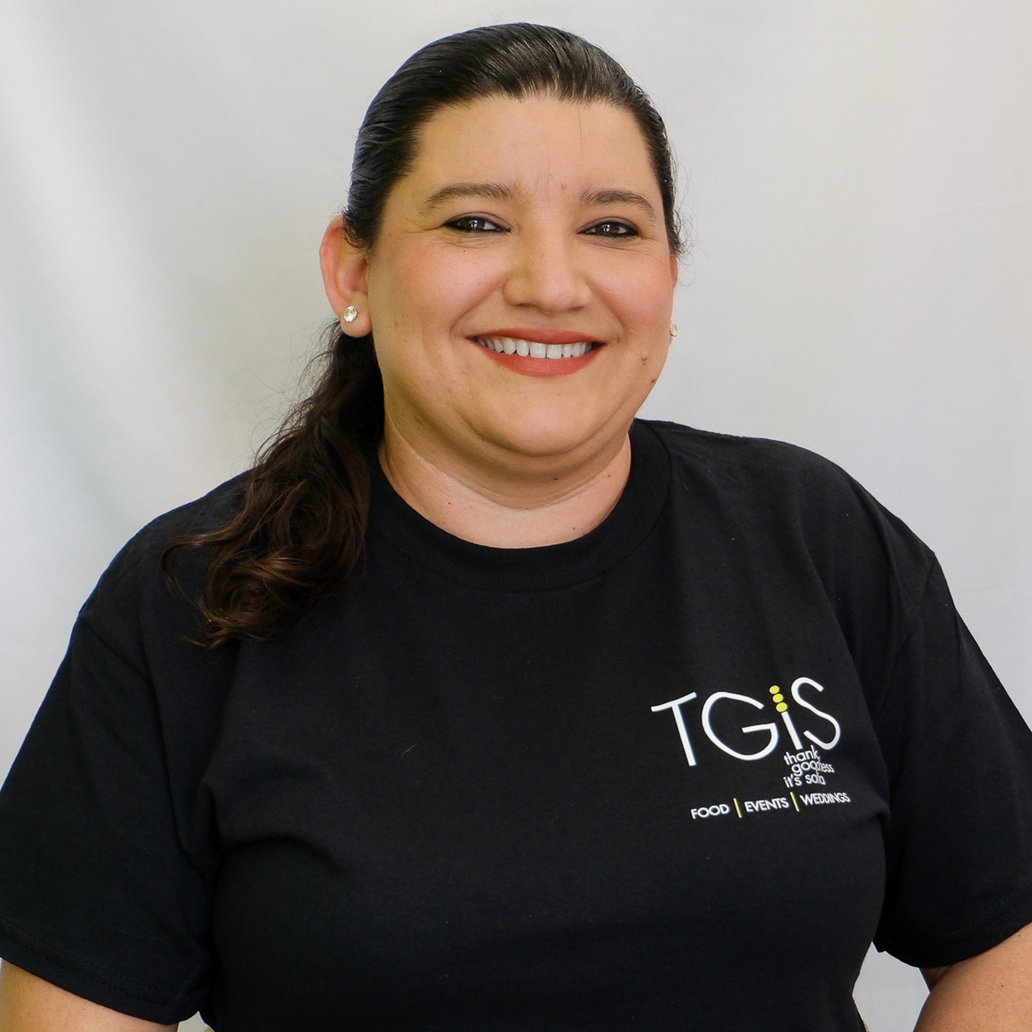 A woman with dark, curly hair smiling with her teeth showing, wearing a black top, against a gray background.