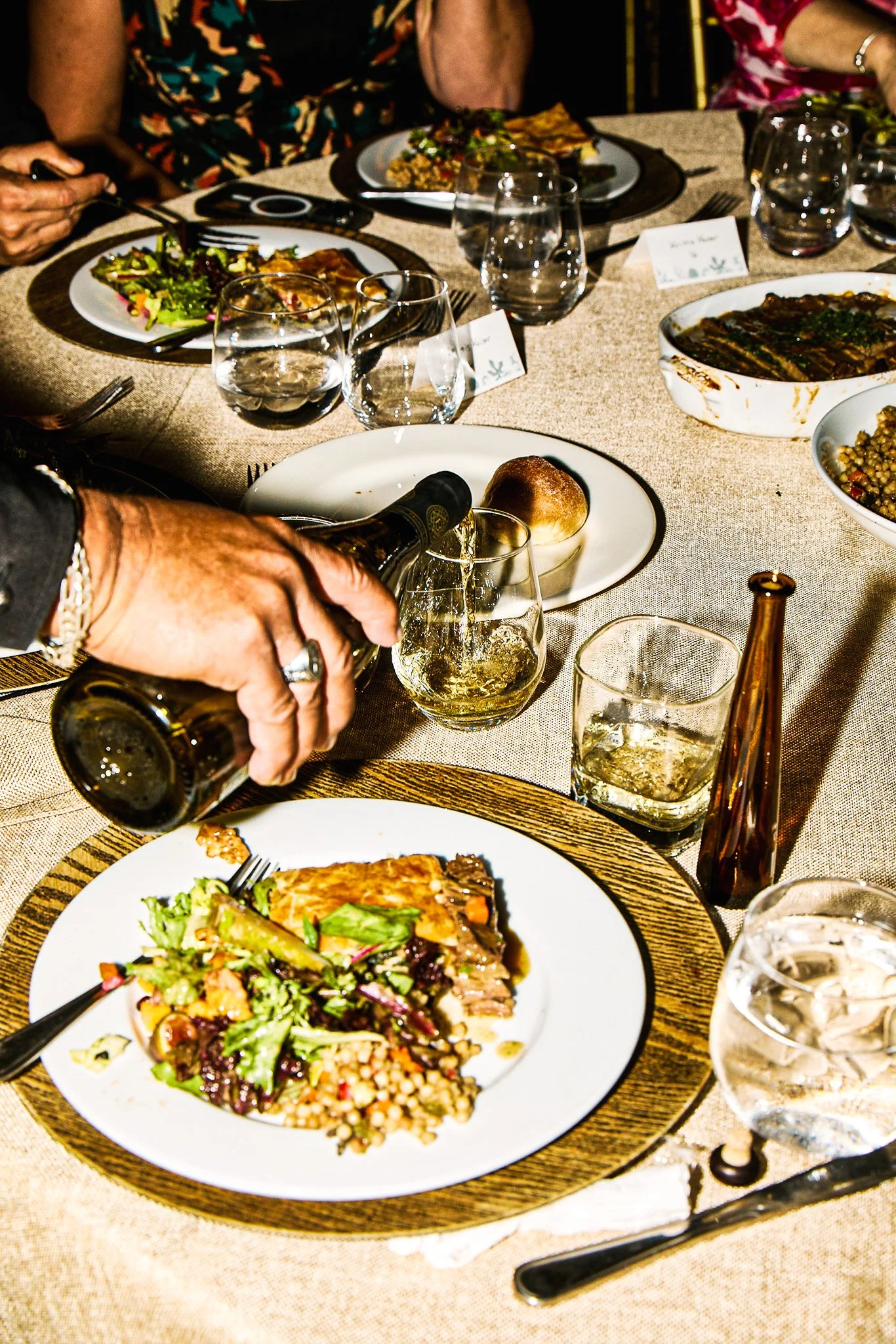 Person pouring white wine into a glass at a dining table with various dishes, glasses, and utensils.