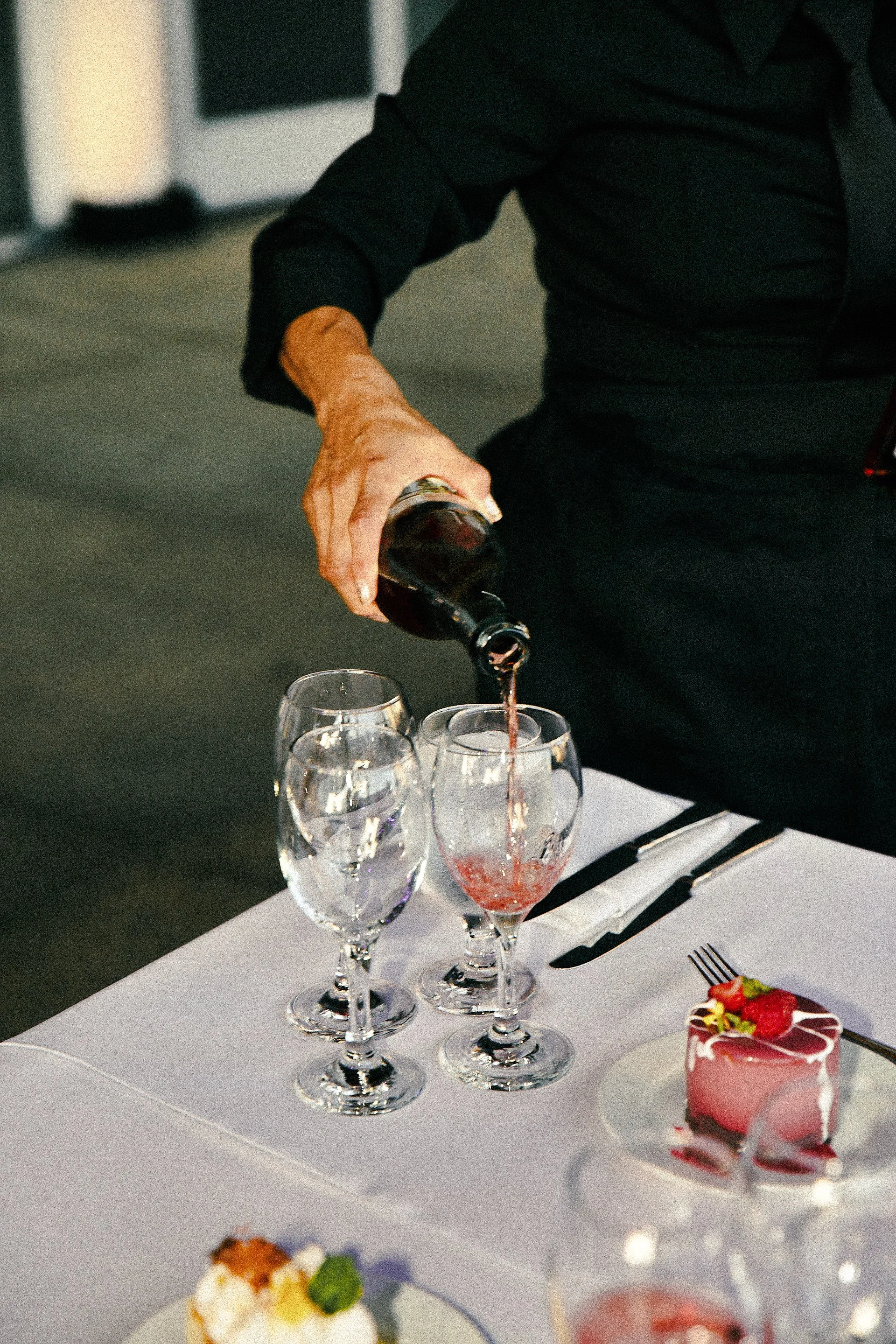 Person pouring red wine into a glass at a table set with glasses, a dessert pink cake with strawberries, and plates of food.