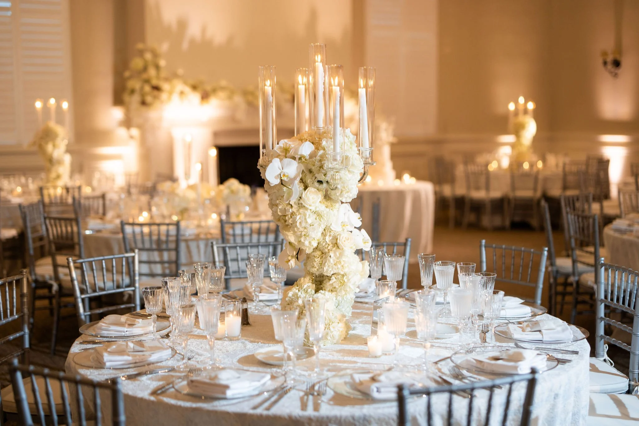 Elegant banquet hall decorated for a wedding reception with round tables, silver chairs, and white floral centerpieces with candles.