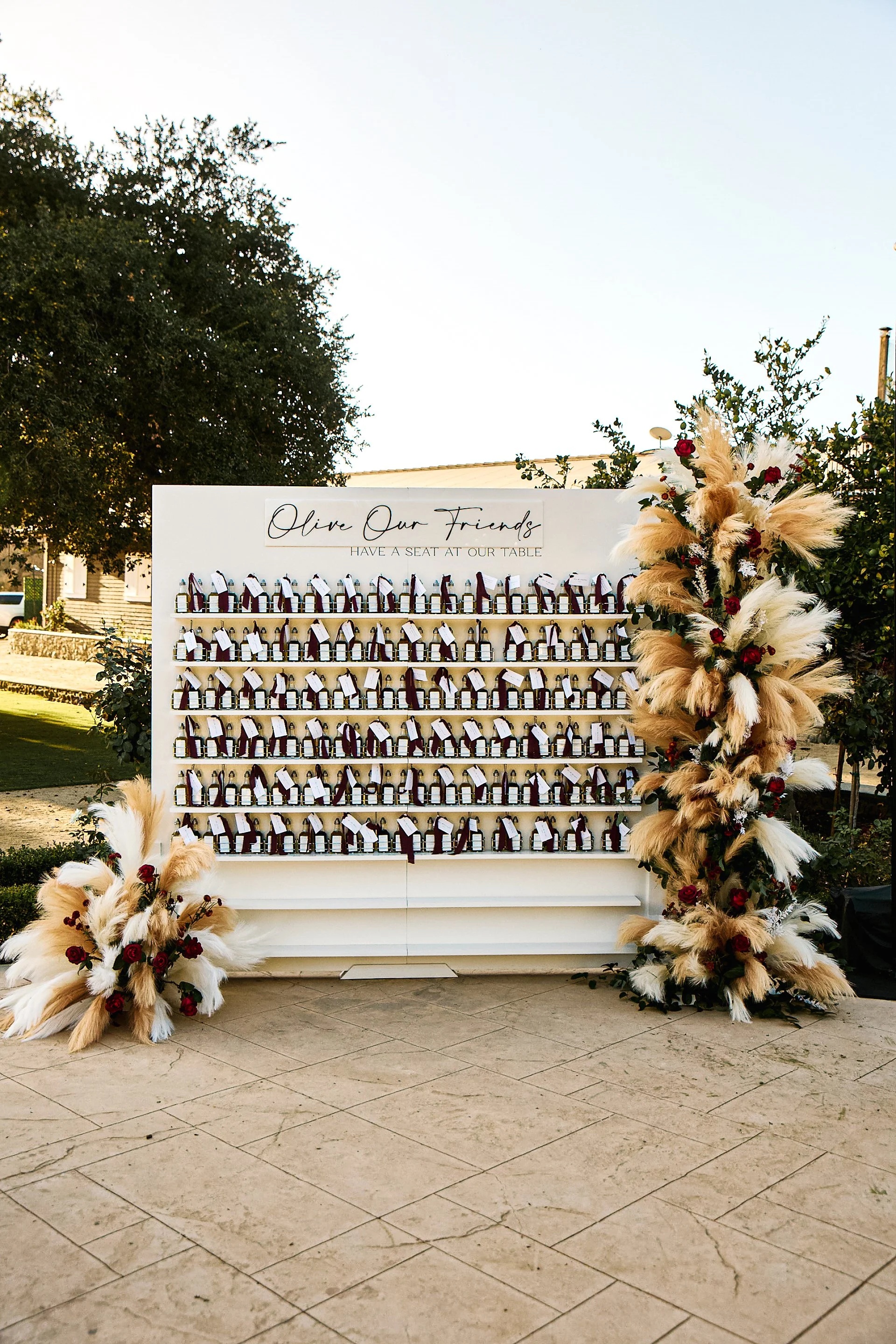 A white display board with bottles, topped with a sign that reads 'Olive Our Friends' and 'Have a Seat at Our Table.' The display is decorated with large floral arrangements of pampas grass, red roses, and greenery on both sides. The scene is outdoor