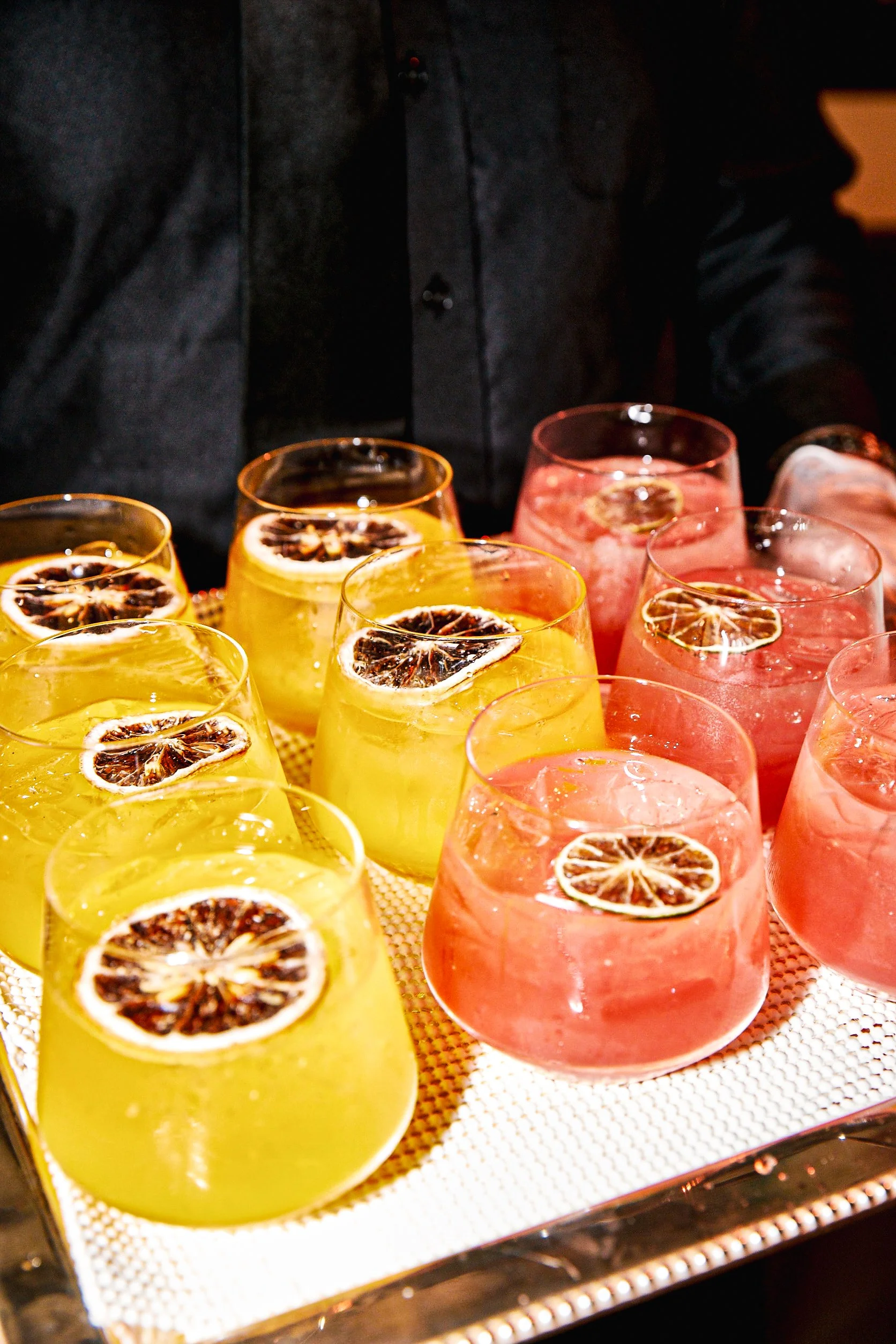 Several glasses of colorful drinks with dried lemon slices on a silver tray, with a person in dark clothing in the background.