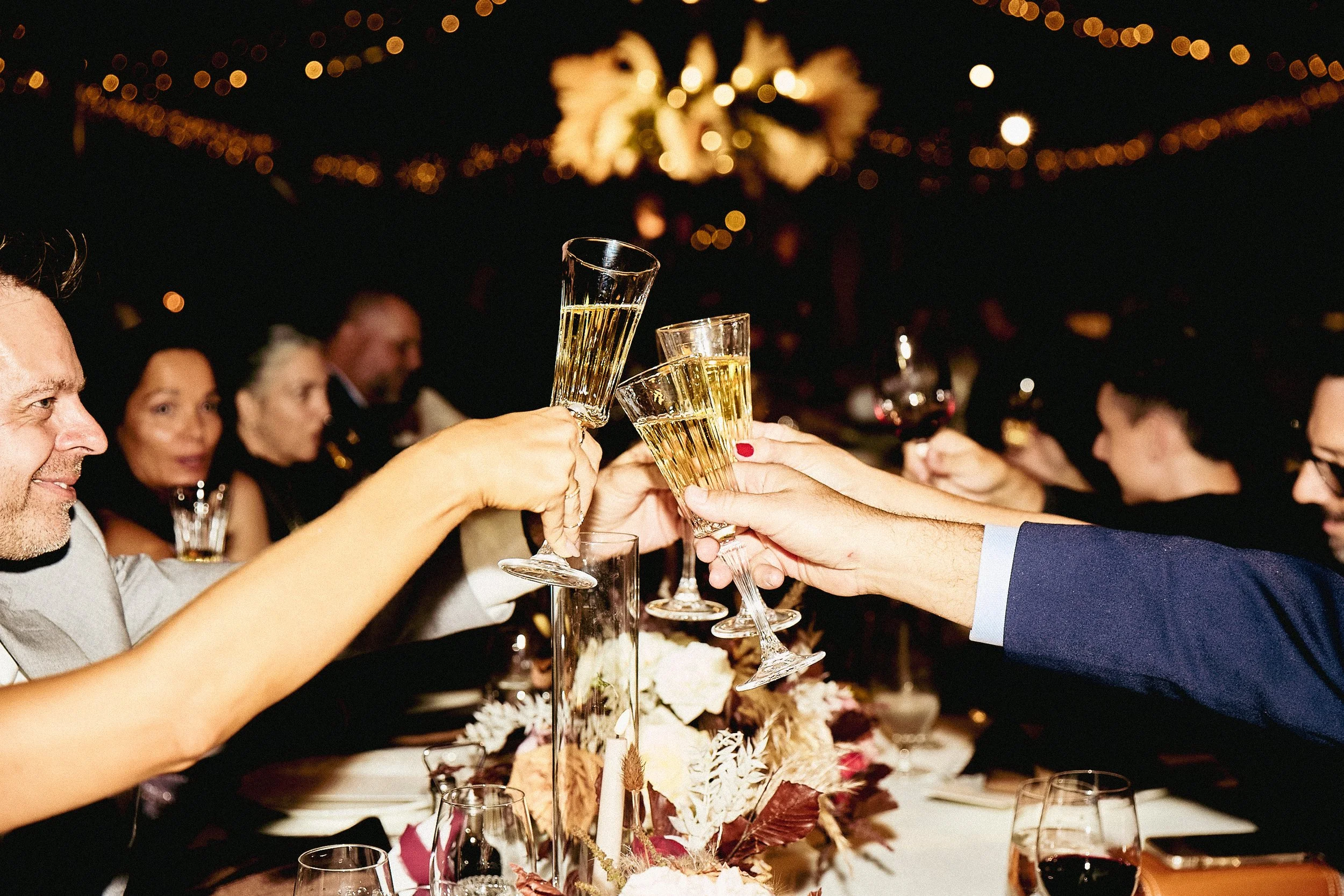 People at a formal event raising glasses of champagne in a toast, with a floral centerpiece on the table and warm lighting decorations in the background.