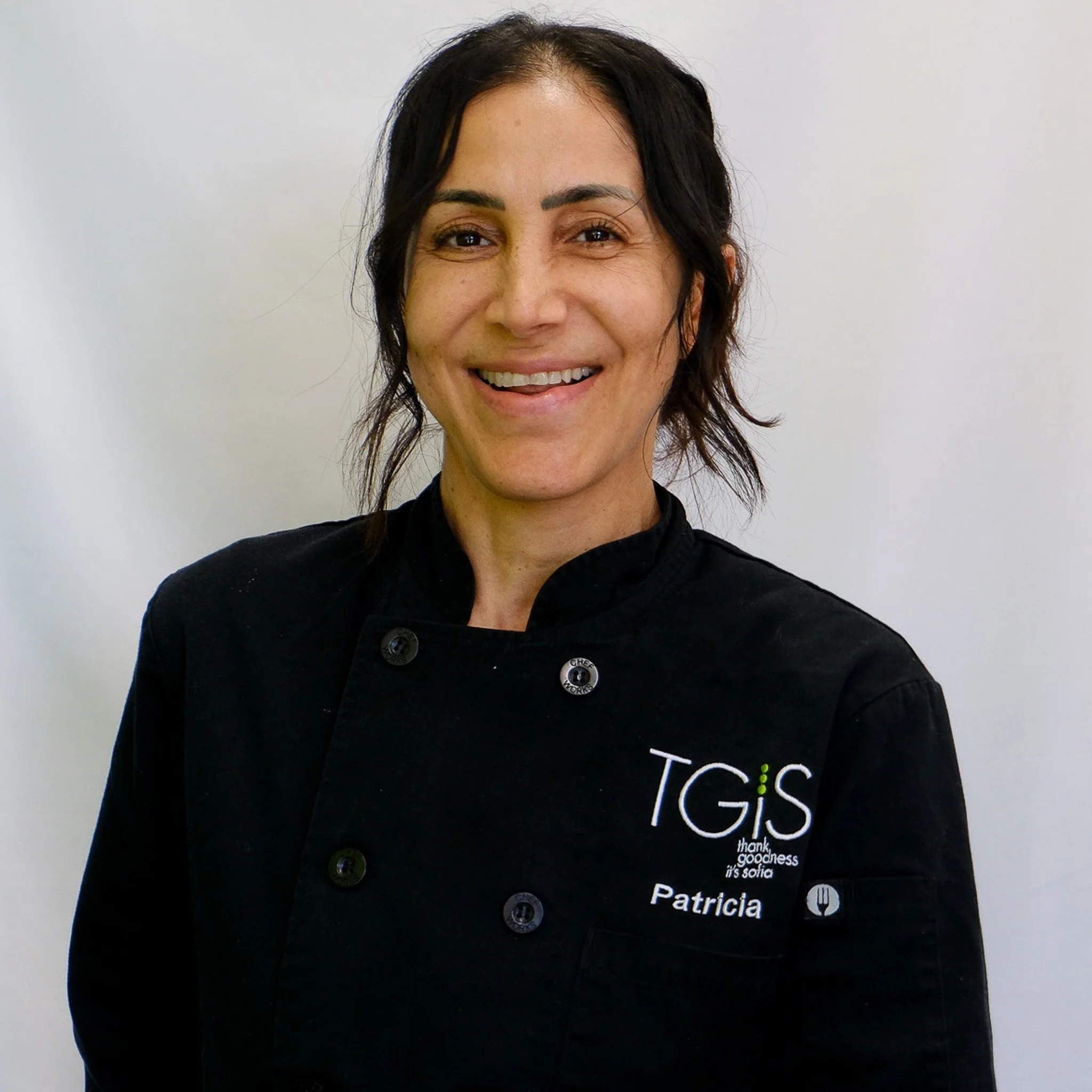 A smiling woman with dark hair in a braid, wearing a black chef's coat with the logo 'TGiS' on it, and a gold cross necklace, standing against a gray background.