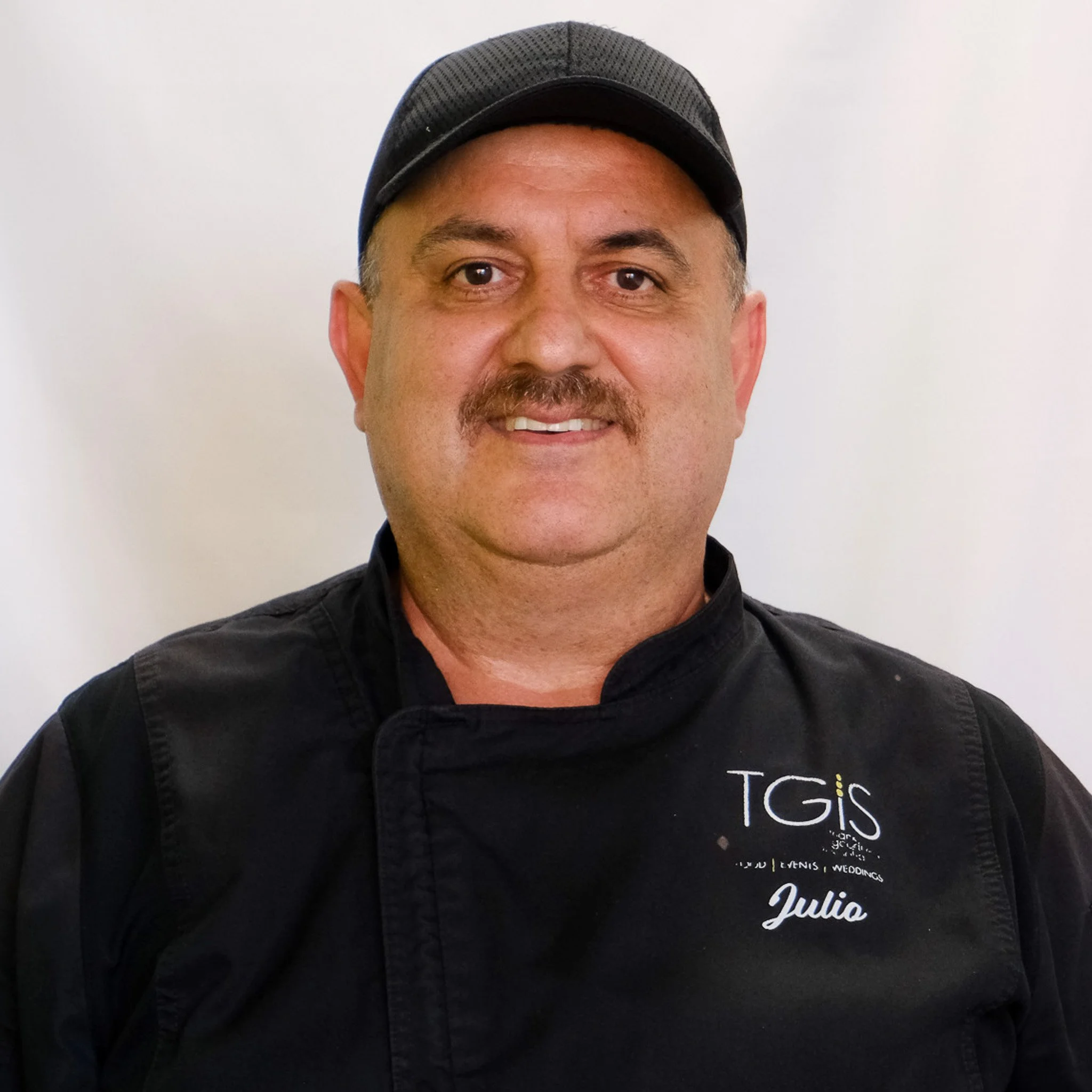 A man wearing a denim cap and a black chef uniform with the logo 'TGiS' smiling at the camera against a gray background.