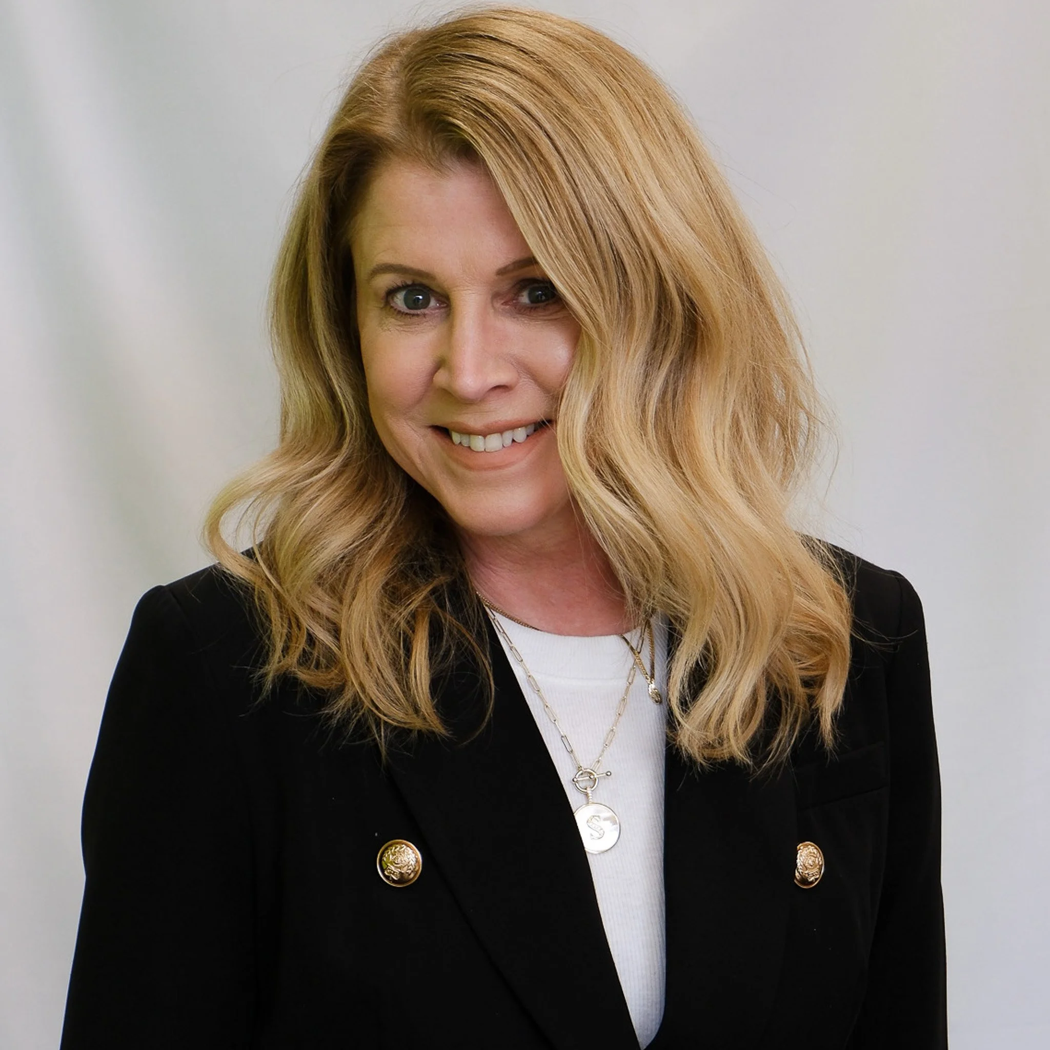 Close-up of a smiling woman with blonde hair wearing a blue blouse and a silver necklace, against a grey background.