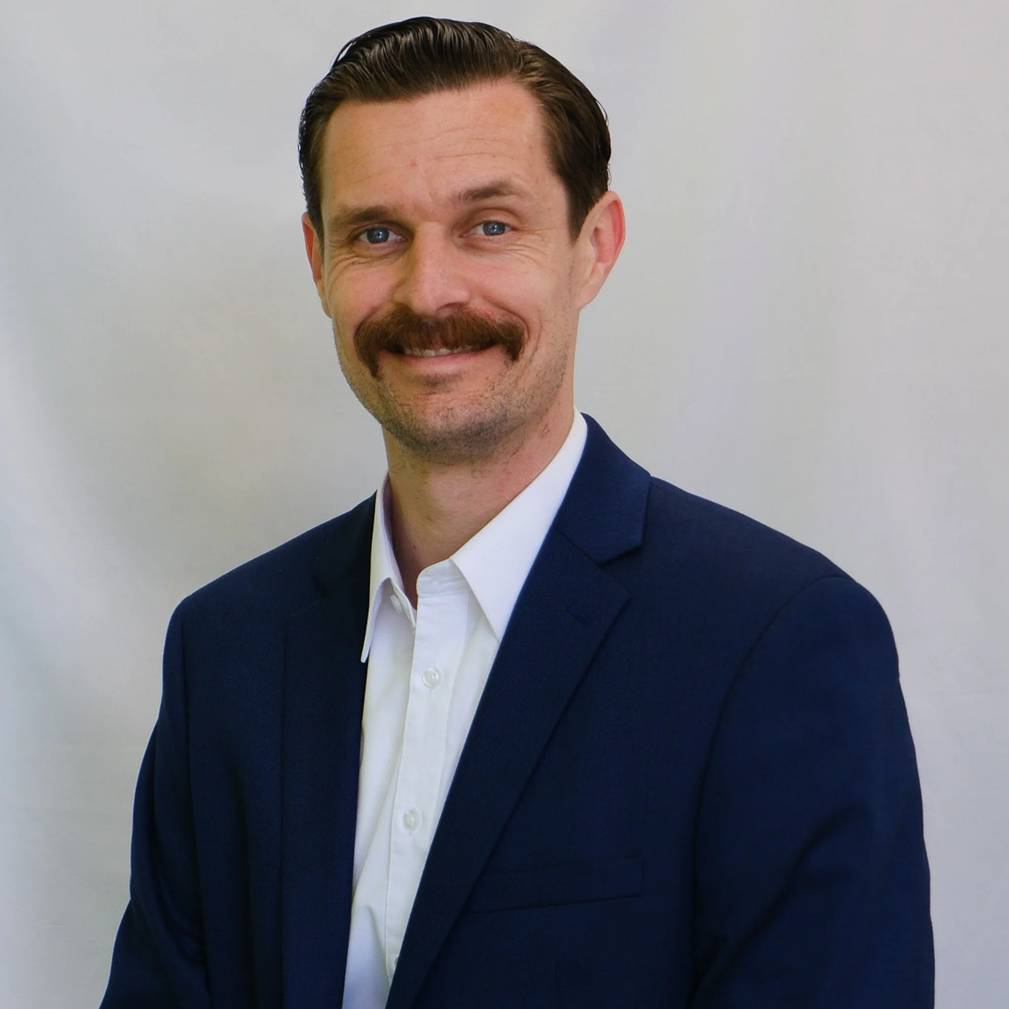 A man with light skin, blue eyes, a large mustache, and wavy brown hair, smiling at the camera against a gray background.