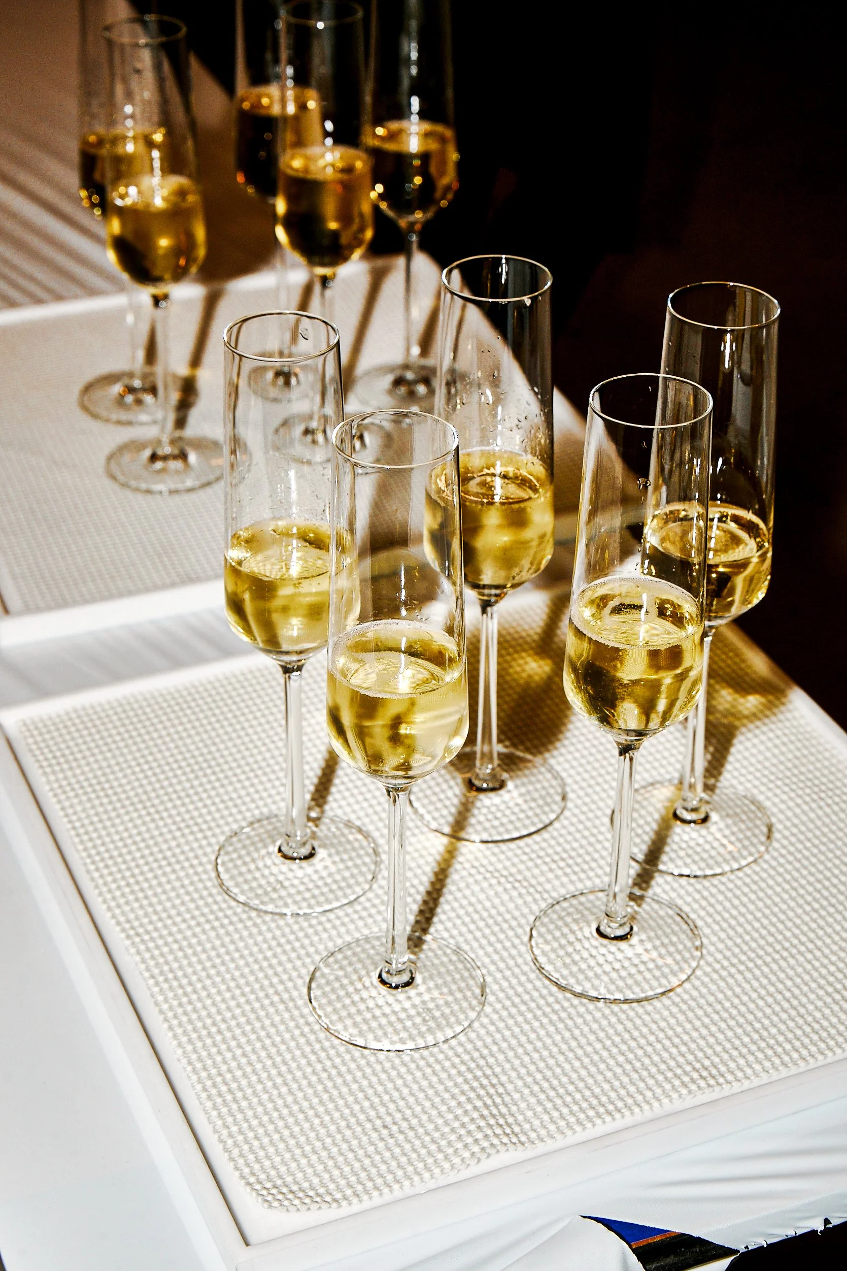 Eight champagne flutes filled with sparkling white wine on a white textured table mat.