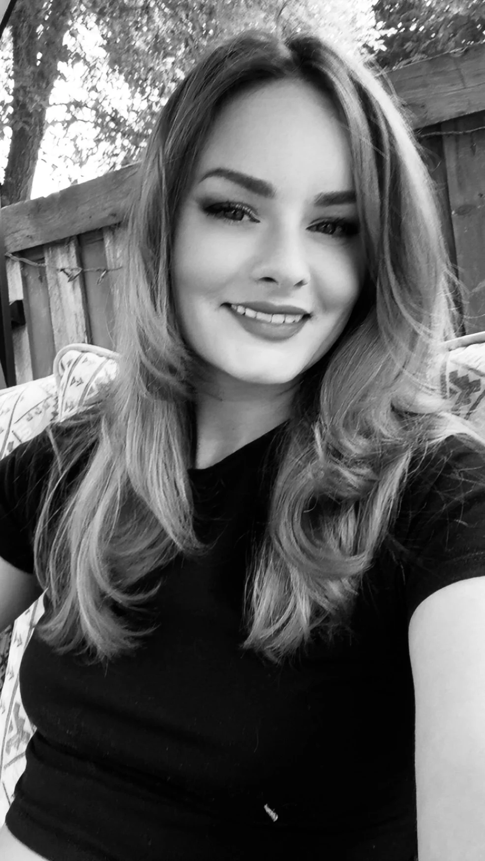 A smiling woman with long, wavy hair taking a selfie outdoors, with a wooden fence and trees in the background.