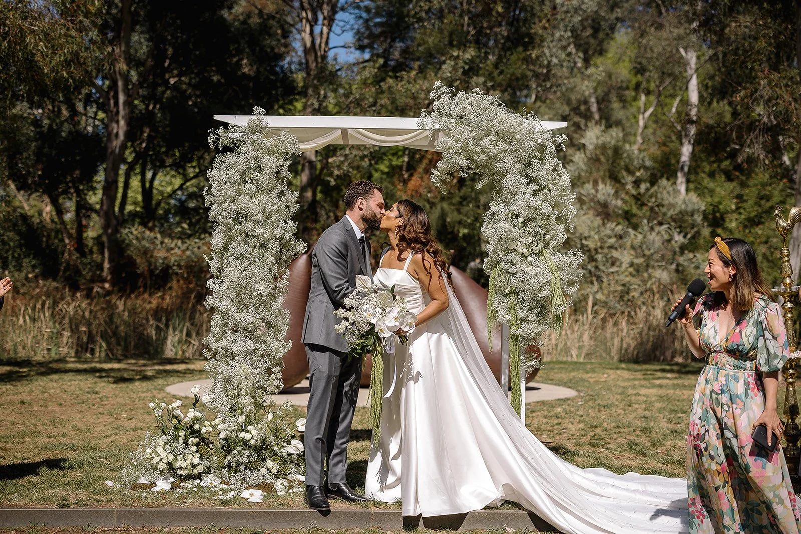 A wedding ceremony outdoors at the National Gallery of Australia with a bride and groom kissing under a floral arch, with a celebrant on the right.