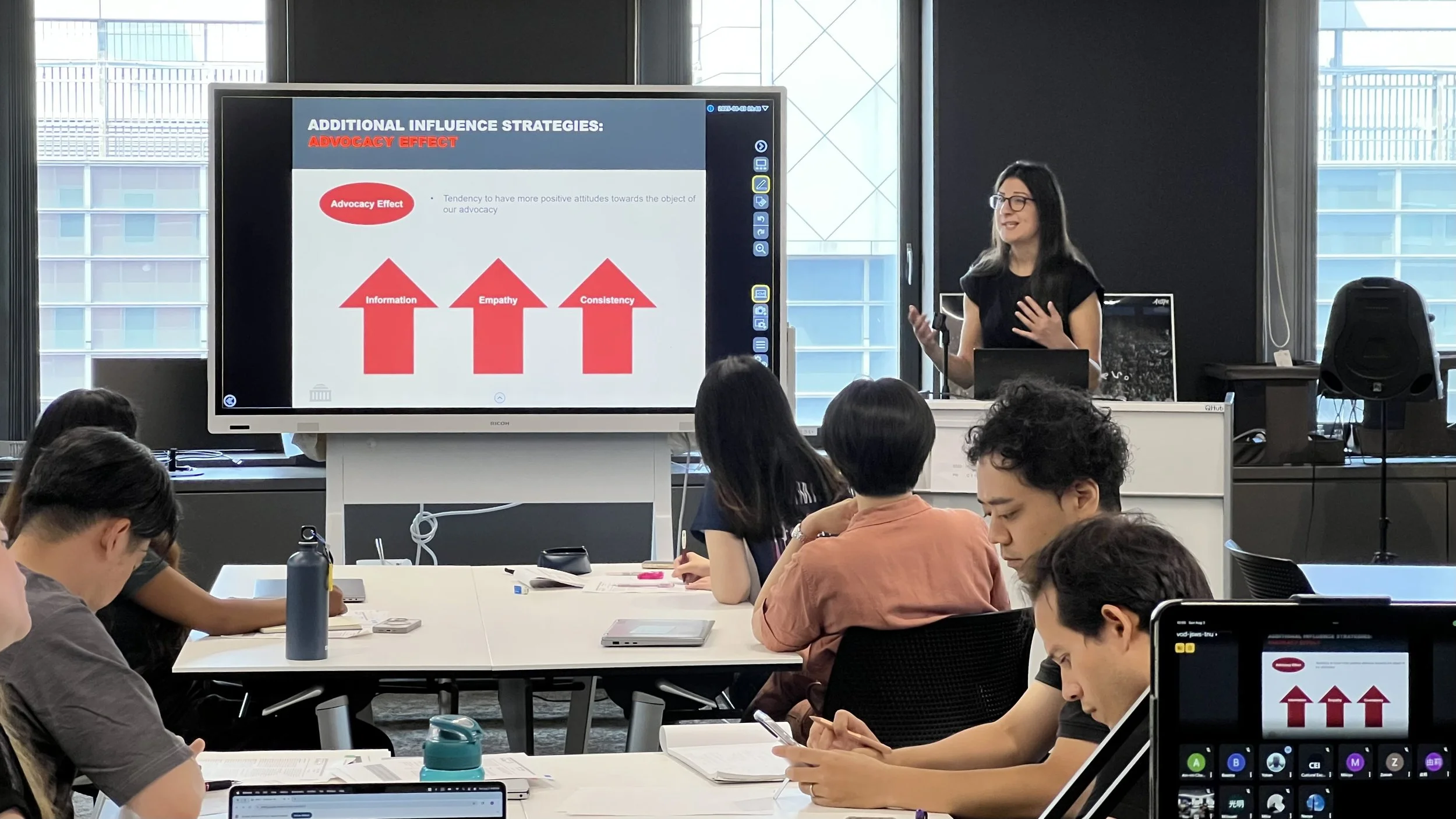 A woman in a black shirt presenting a slide on a large screen during a classroom or seminar, with students taking notes and using electronic devices.