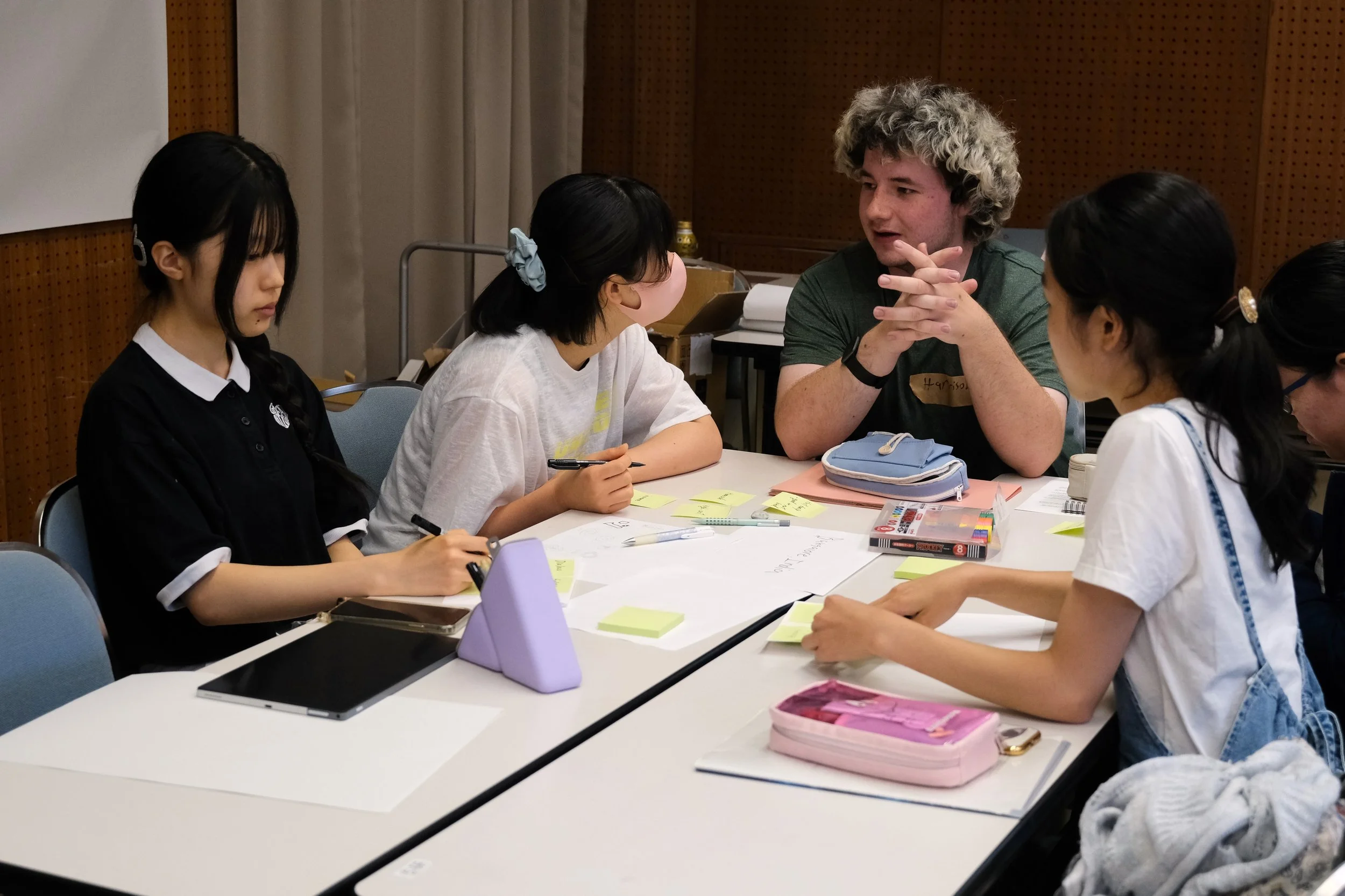 Group of five young women and a young man sitting around a white table engaged in discussion with notes, pens, tablets, and stationery on the table.