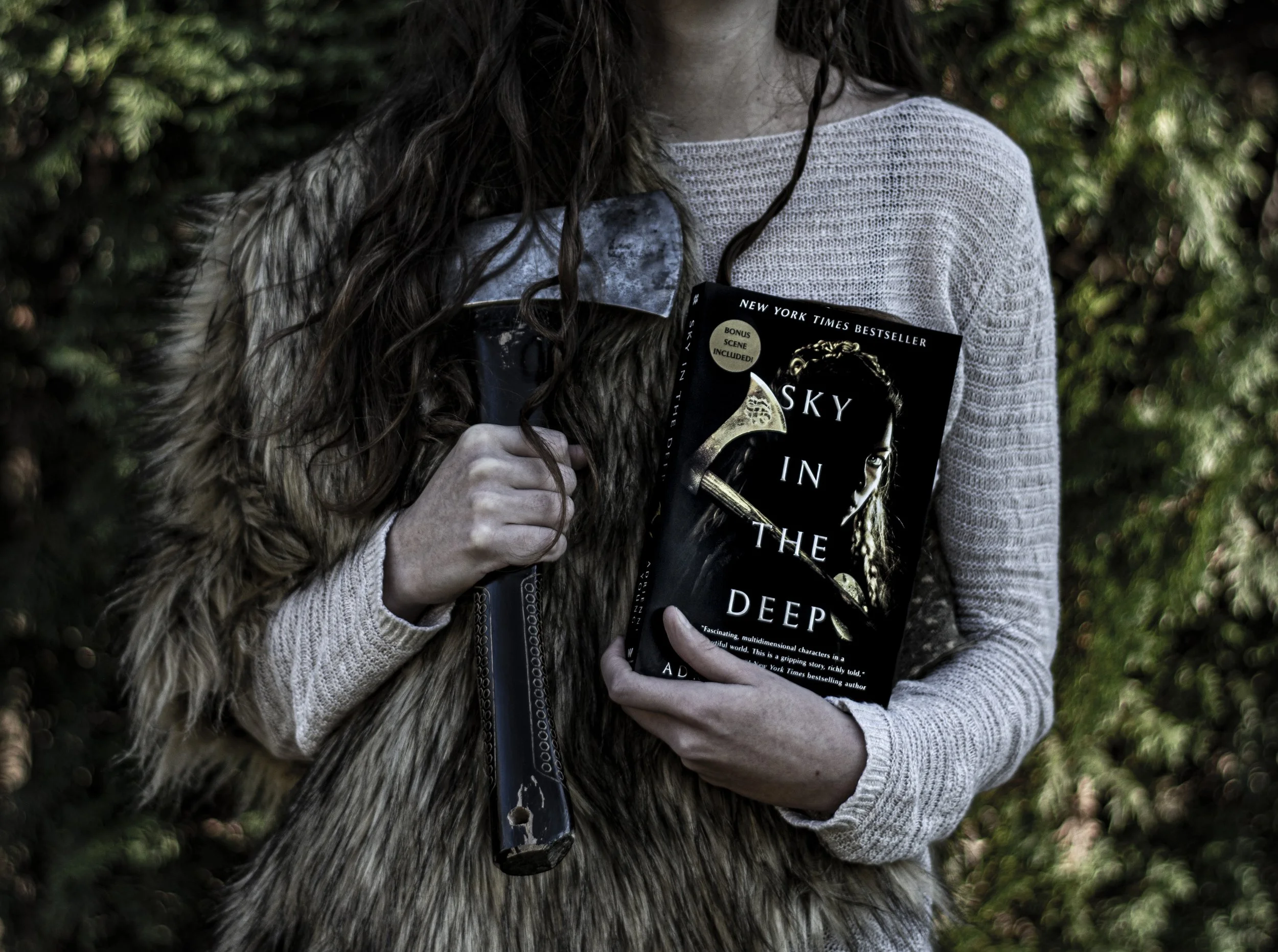 Eve stands in front of a wooded area wearing a fur pelt. In one hand she holds an axe. In the other hand she holds the book Sky in the Deep by Adrienne Young.