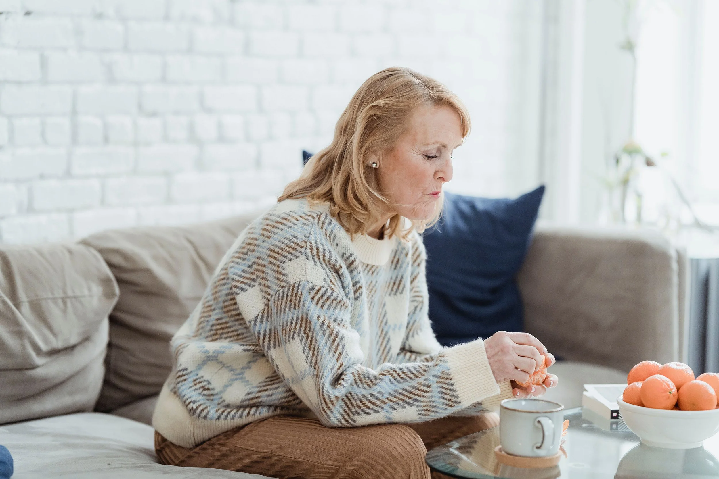 A woman in a patterned jumper reaches for a mug of tea on the coffee table in a bright living room.