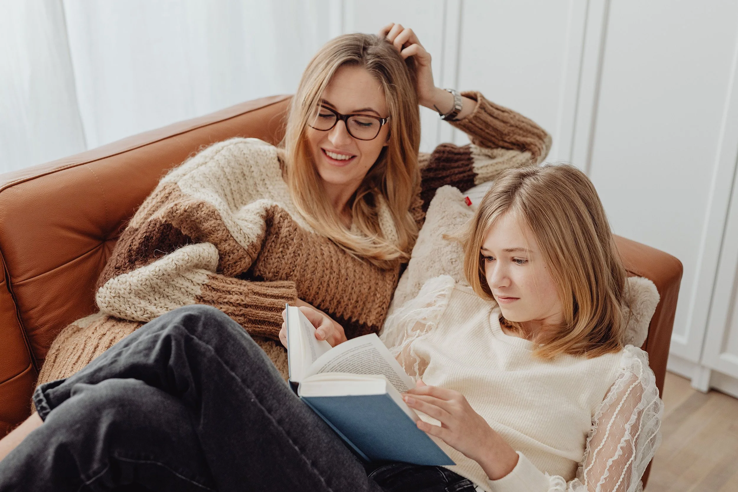 A woman in glasses smiles as she sits on a leather sofa beside a teenage girl reading a book.