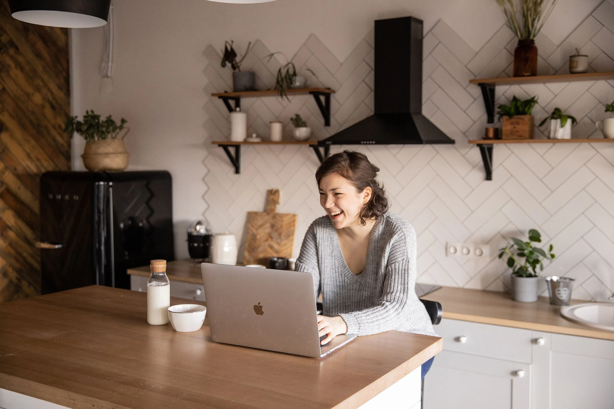 A woman laughs while working at her laptop at a kitchen bench in a bright, plant-filled home.