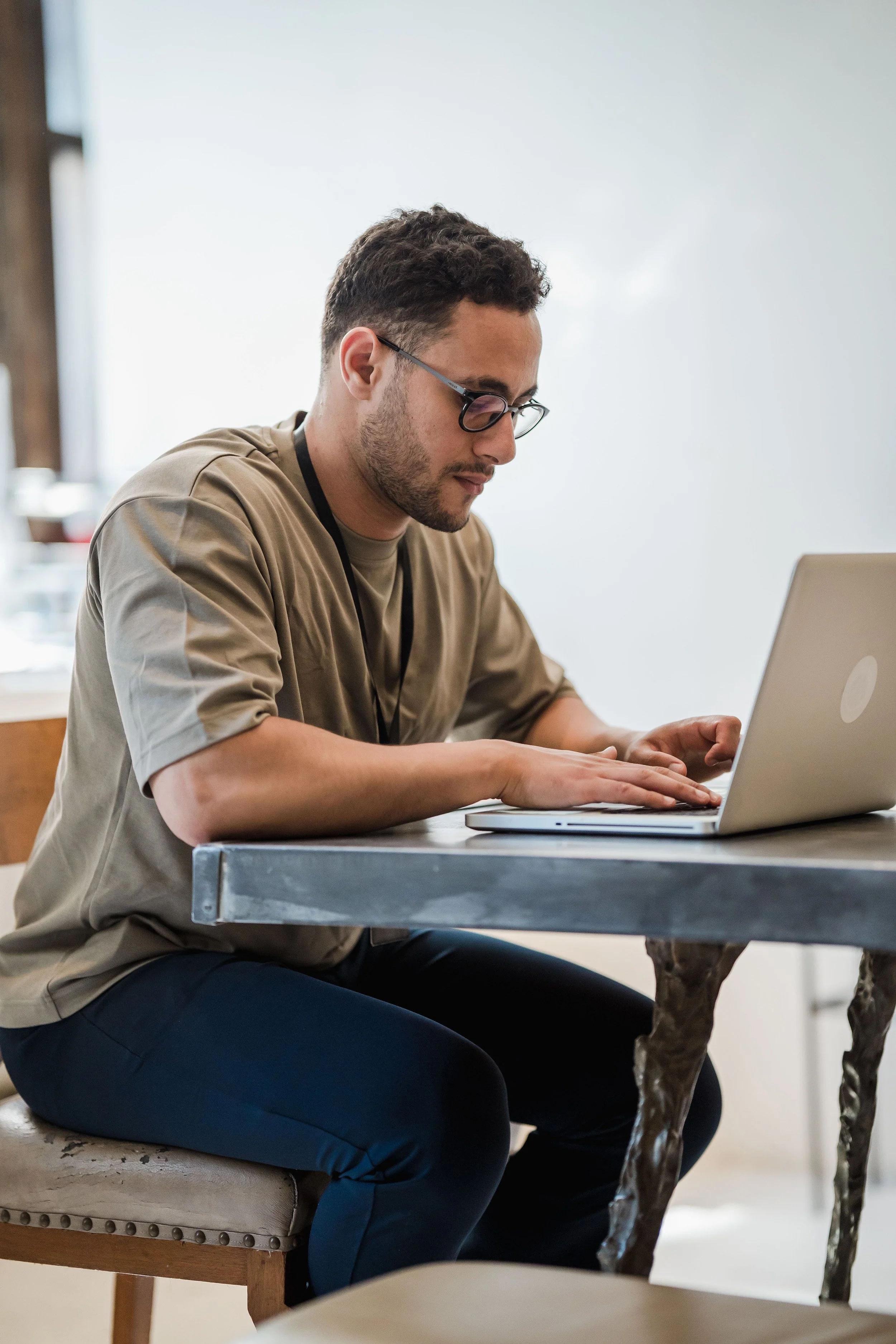 A man in glasses types at a laptop at a table in a bright, uncluttered room.