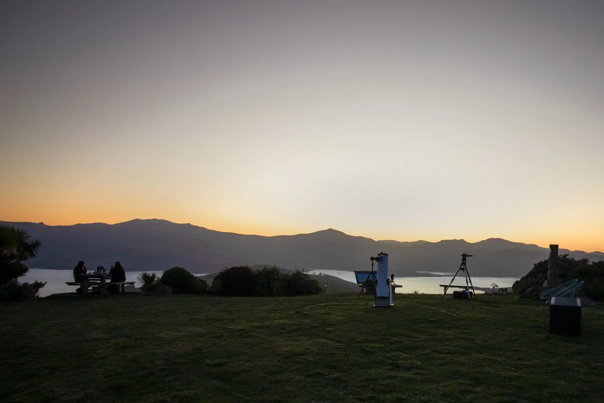 elescopes and a picnic table set up on a grassy hill overlooking Akaroa harbour at sunset, ready for stargazing.