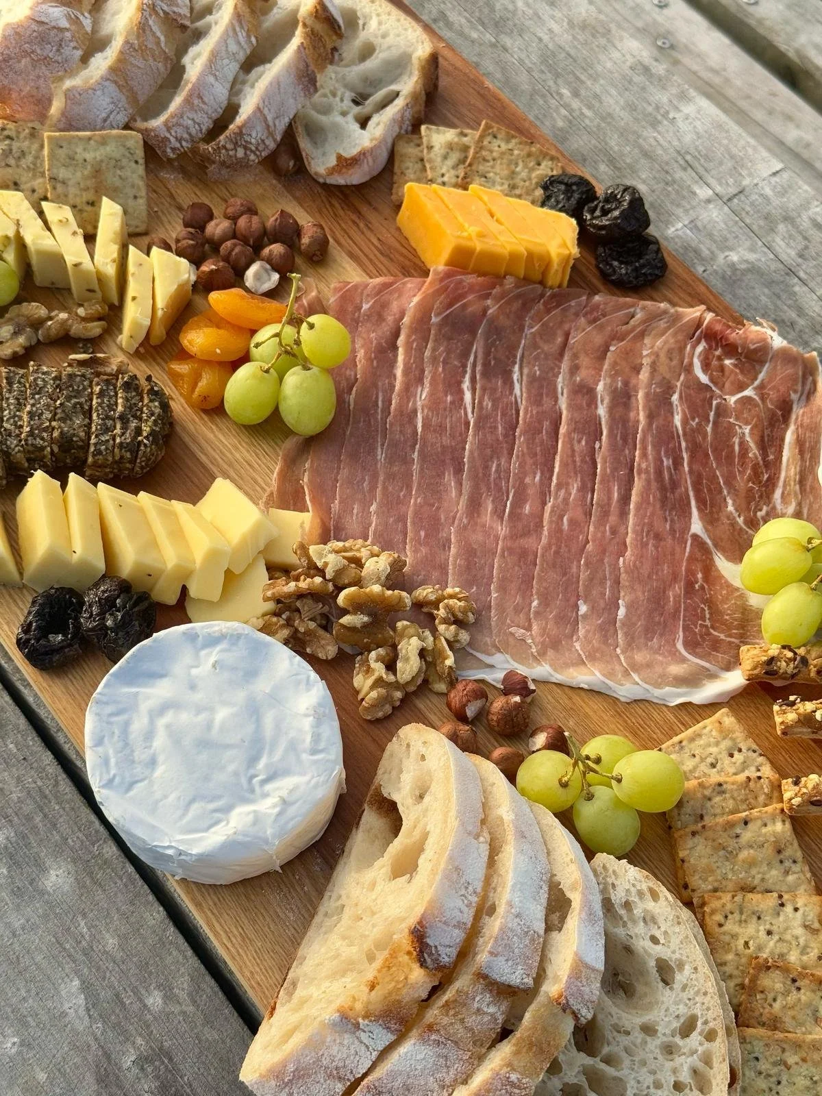 wo people seated at a wooden picnic table with a charcuterie grazing platter and drinks, pointing towards a scenic Akaroa harbour and hills at dusk.