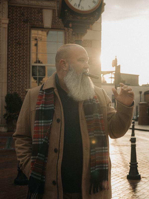 A man with a bald head and a long gray beard is standing outdoors in front of a brick building with a clock. He is wearing a beige coat and a colorful plaid scarf, holding a cigar in his mouth, and making a gesture with his hand as the sun sets in the background.