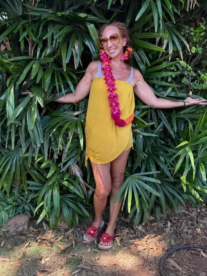 Woman in yellow dress and flip-flops with pink flower accessories standing among lush green tropical plants.