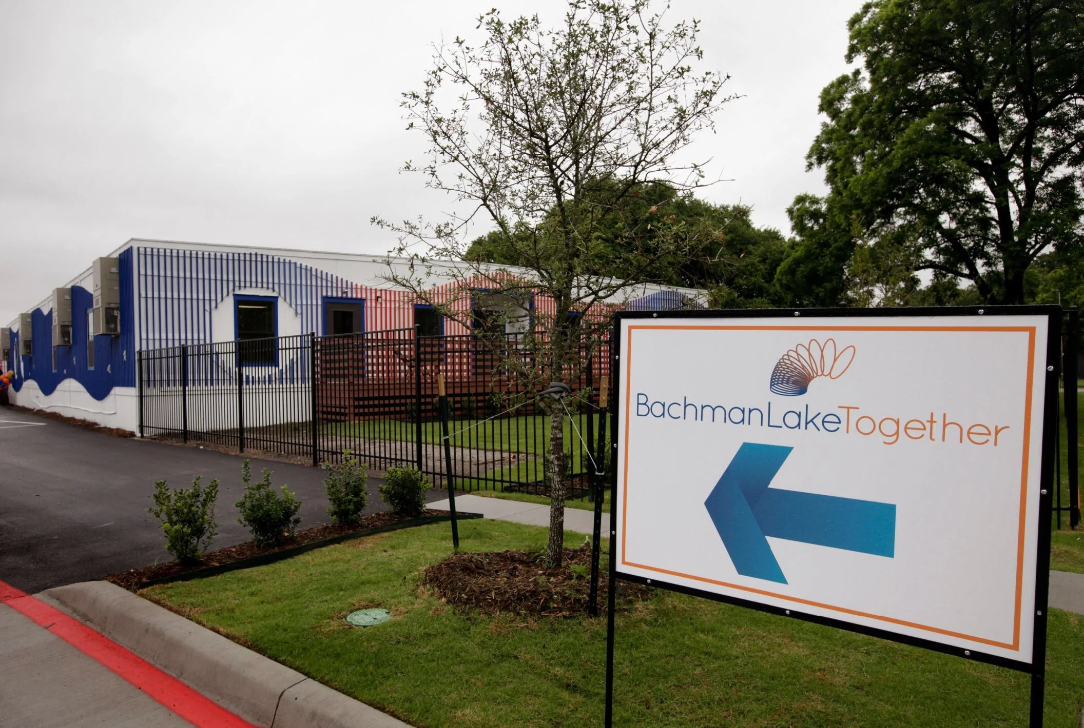 Sign for Bachman Lake Together with a blue arrow pointing left, outside a building with colorful wall art and a small tree nearby.