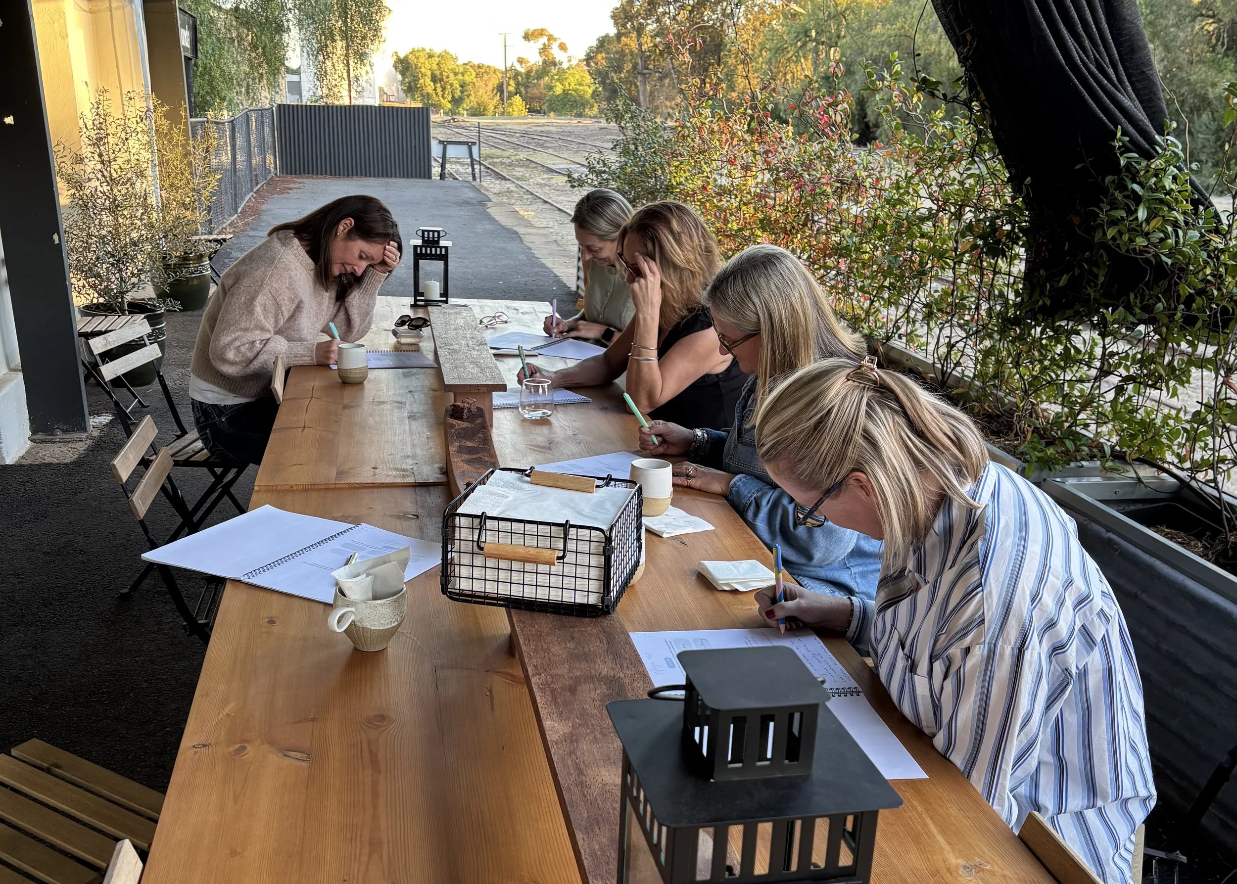 Six women sitting at a long outdoor wooden table, focused on writing or reading documents, with glasses and notebooks, sitting under a canopy with plants and train tracks visible in the background.