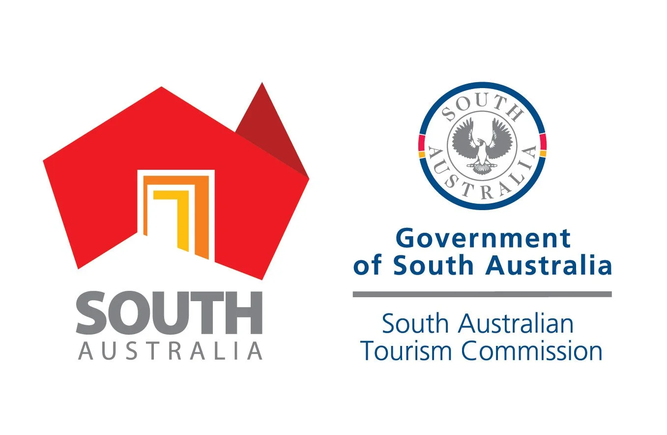 Logo of South Australia Tourism Commission with government emblem, featuring a stylized red house and a circular emblem of a black swan, with text indicating the Government of South Australia and South Australian Tourism Commission.