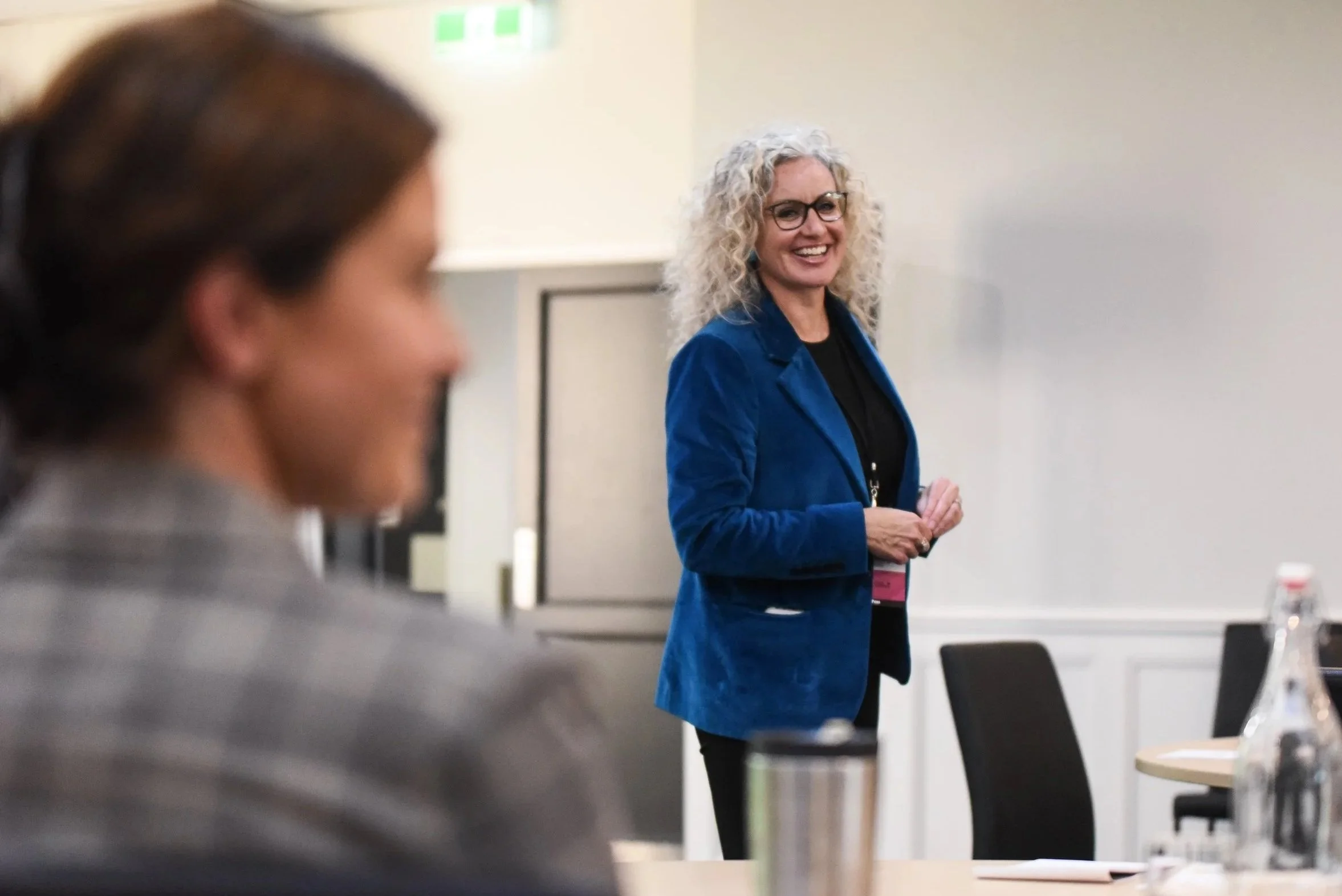 A woman with curly blonde hair, glasses, and a blue blazer smiling as she stands in a conference room, with another woman blurred in the foreground. Lesley Magill.