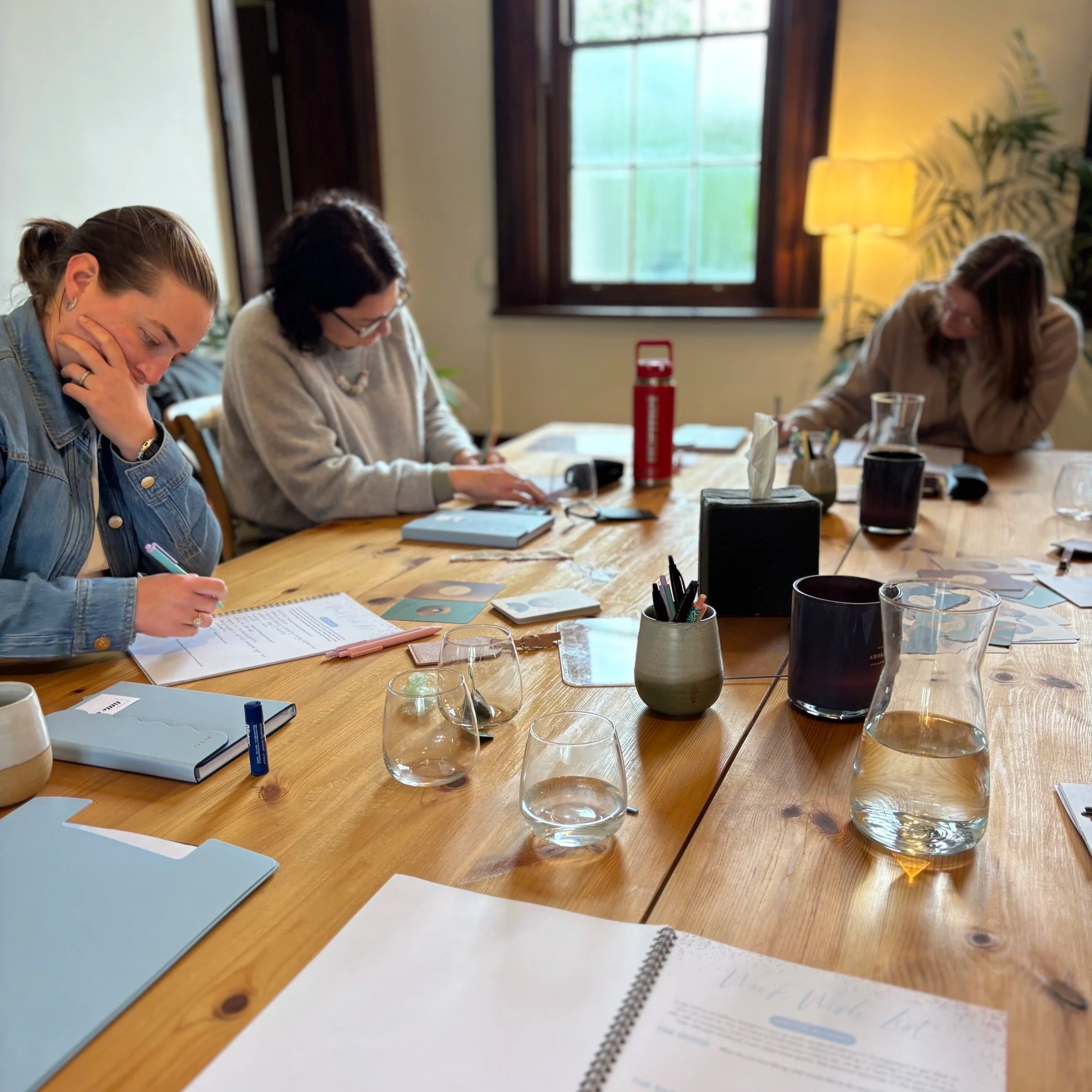 A group of five women sitting at a long outdoor table, engaged in writing or reading, with one woman smiling and laughing, all of them appearing focused and relaxed.