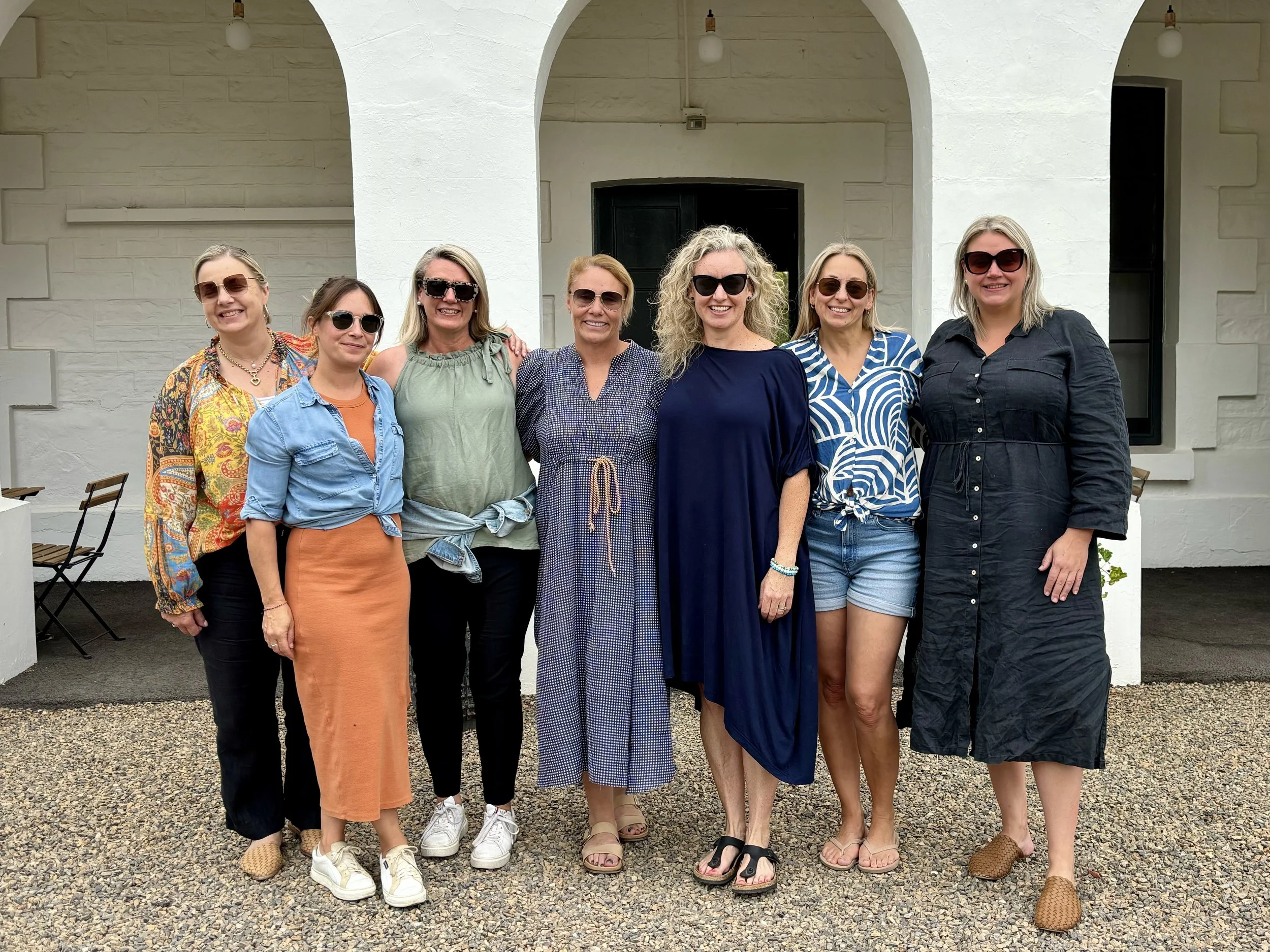 Little Shove Retreat. A group of eight women standing side by side in front of a white building with arched doorways, all smiling and wearing sunglasses.